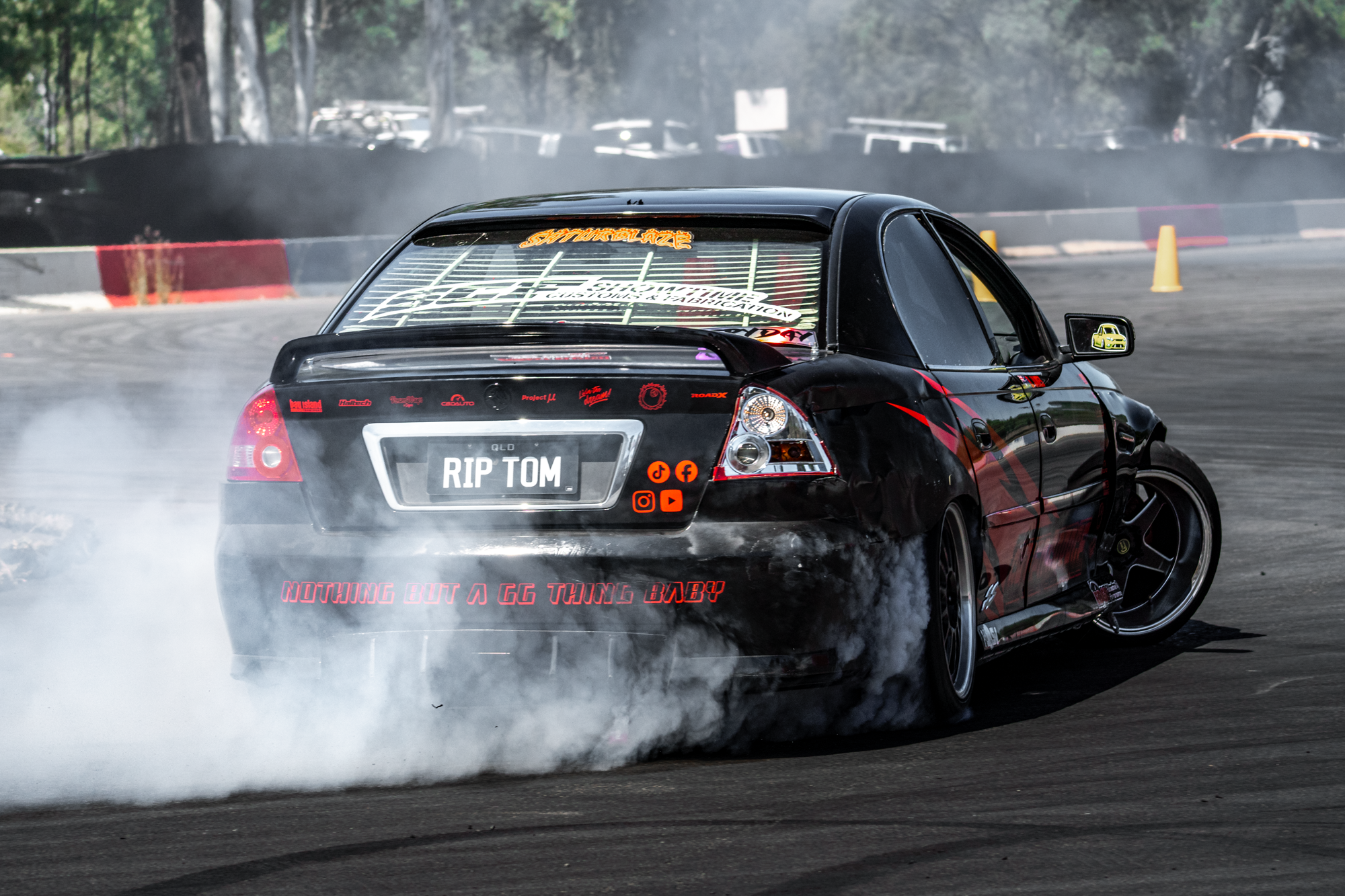 Black car performing a drift on a racetrack, with smoke coming from the tires, during daytime. The car has various decals, a license plate saying 'RIP TOM', and a rear window sticker reading 'SAPPHIRE LAZE'.