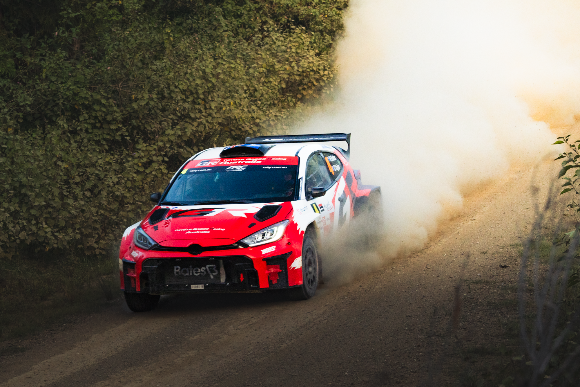 Red and white rally car speeding through a dirt road, kicking up dust, surrounded by green foliage.