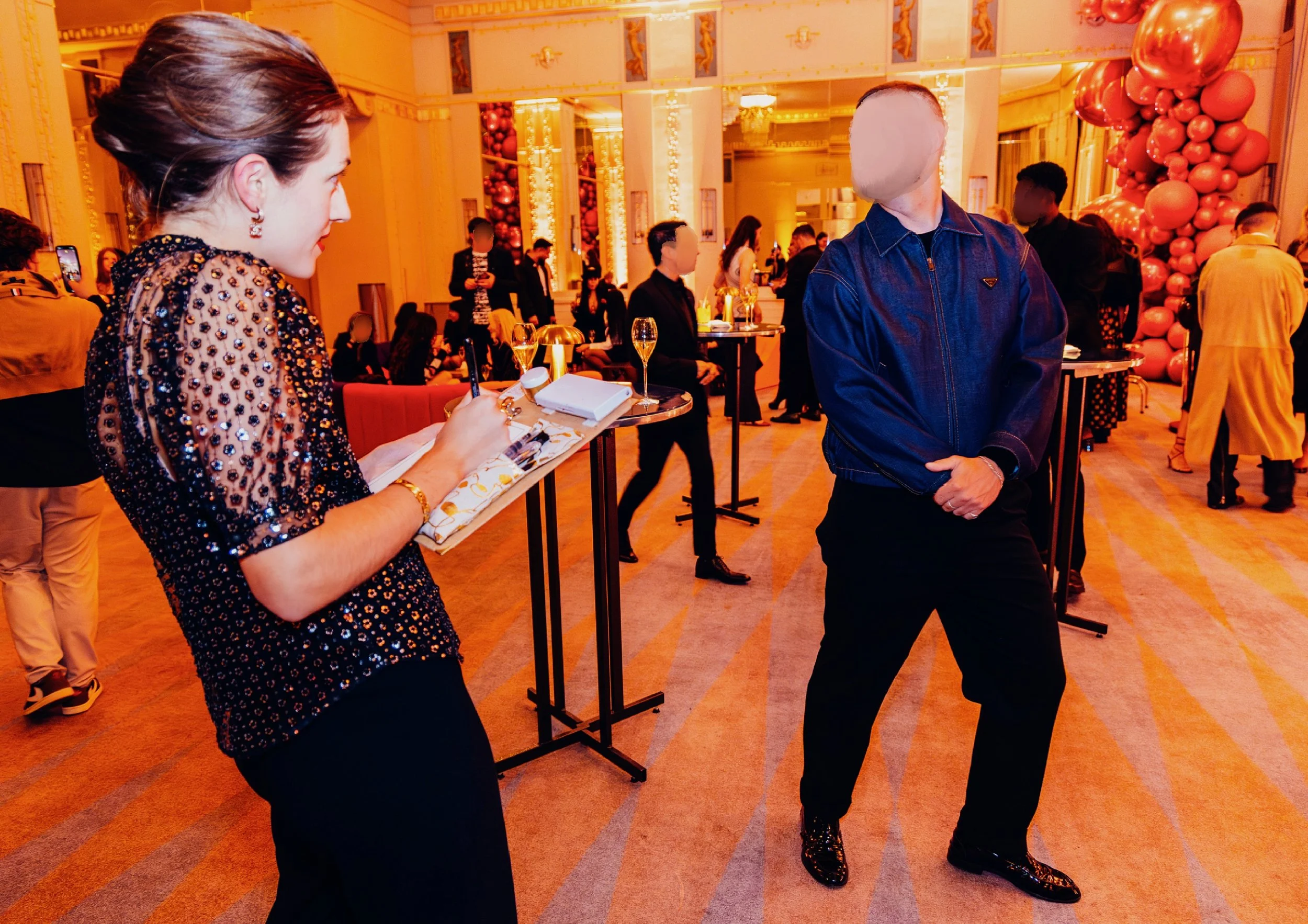 Une femme écrit quelque chose dans un carnet lors d'une soirée élégante. Un homme avec un visage flouté porte une veste en denim noir. La salle est décorée avec des ballons roses et des lumières dorées.