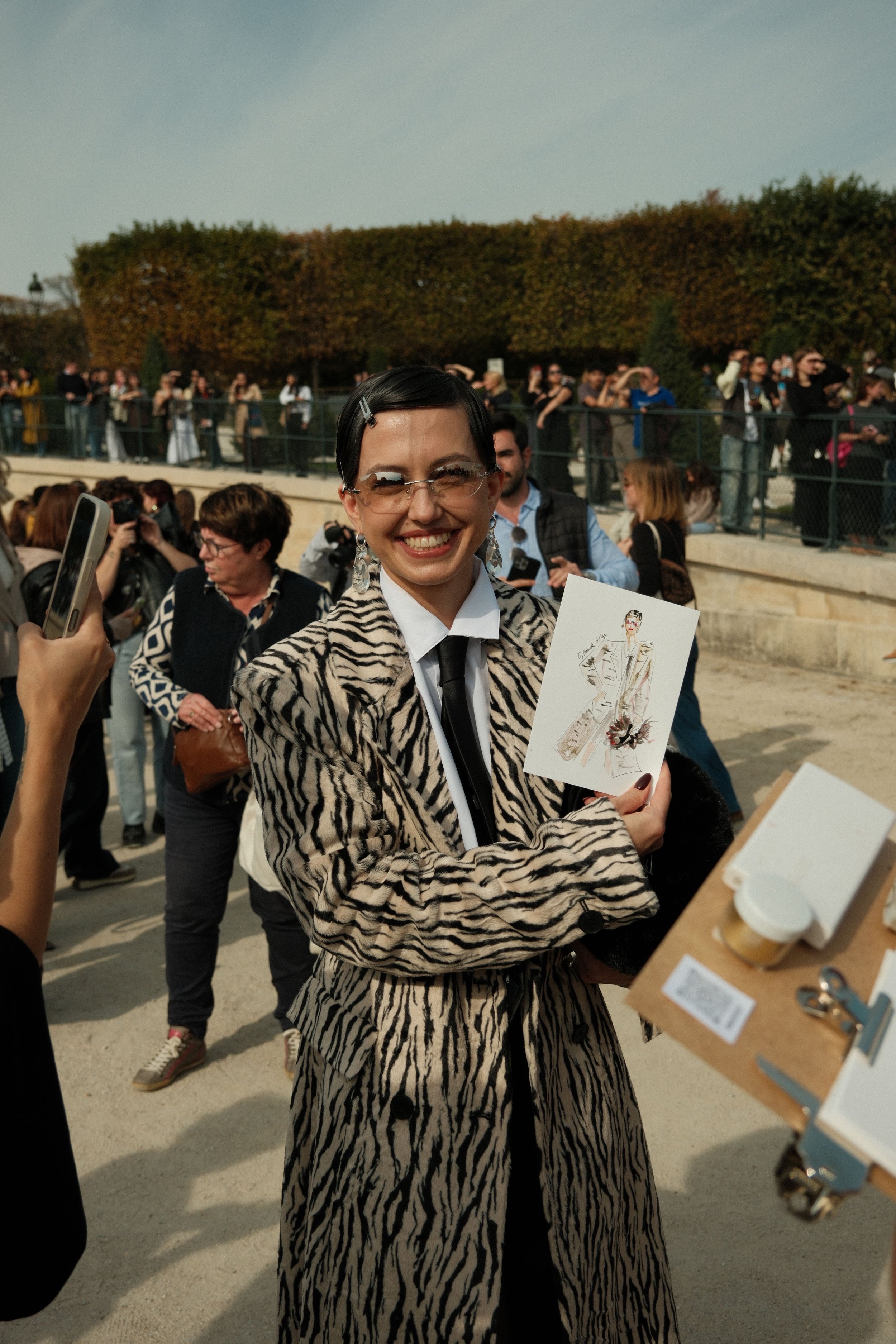 Une femme souriante portant un manteau à motif animal, tenant une carte et un plateau avec une tasse de café, entourée de plusieurs personnes lors d'un événement en plein air.