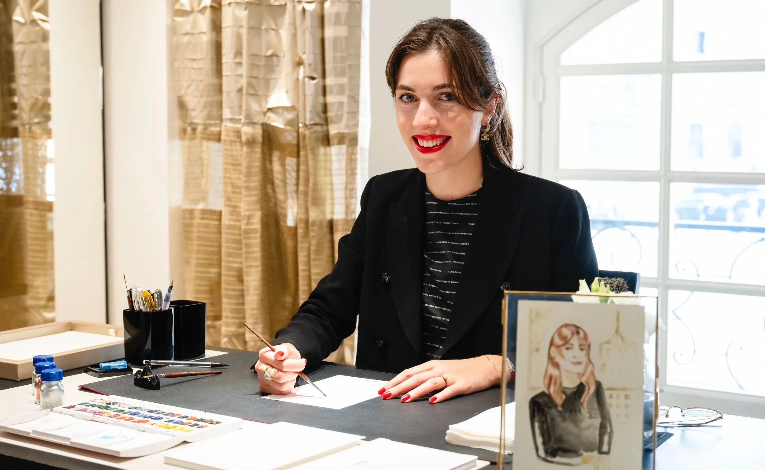 Une femme assise à une table, tenant un pinceau, entourée de matériaux pour la peinture, souriant à la caméra, dans une pièce lumineuse avec de grandes fenêtres. Sur la table, une peinture sur toile et d'autres fournitures artistiques.