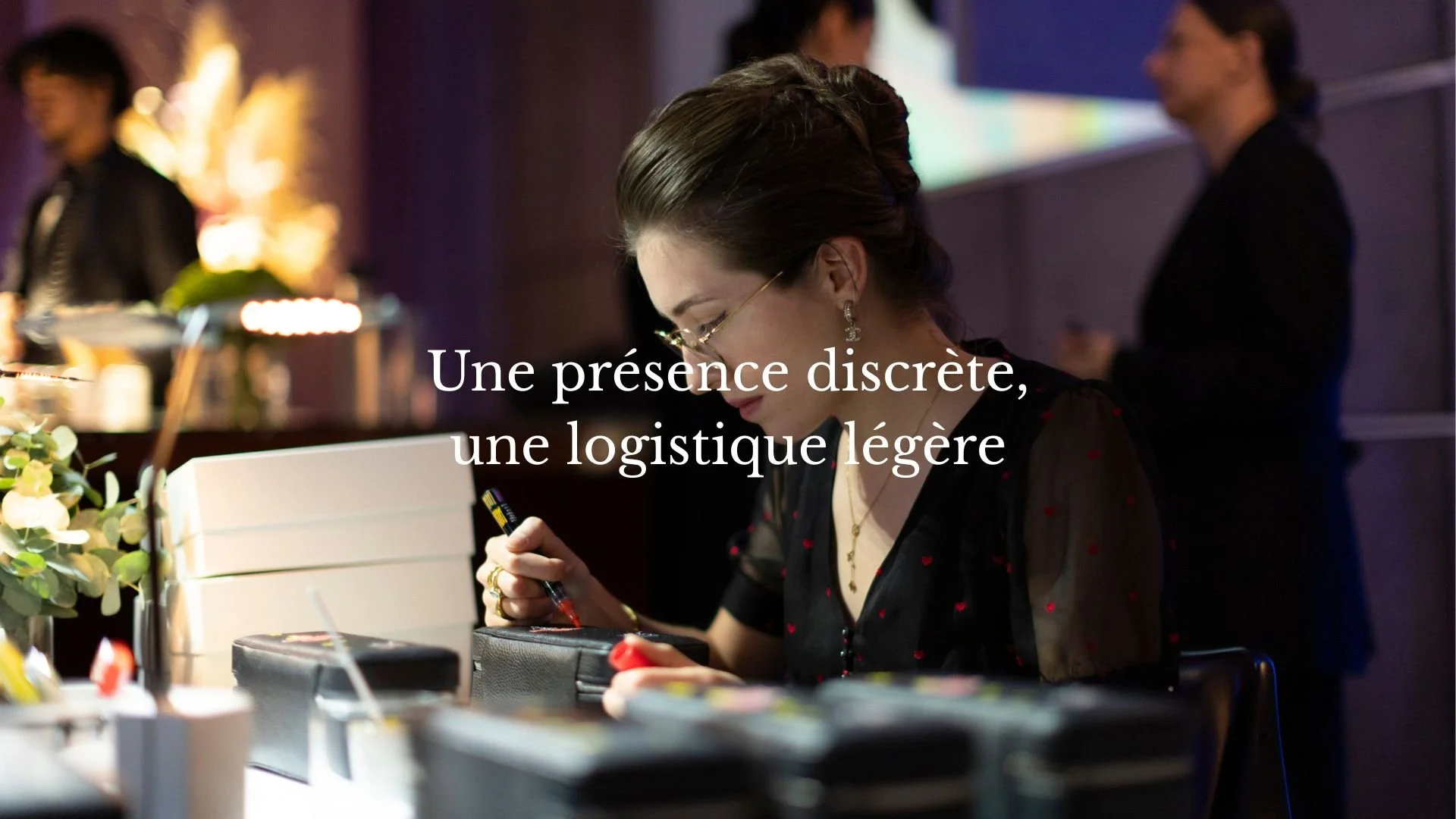 Une femme habillée élégamment regarde un carnet ou un agenda, dans un intérieur moderne avec des lumières tamisées, accompagnée de deux autres personnes floues en arrière-plan.