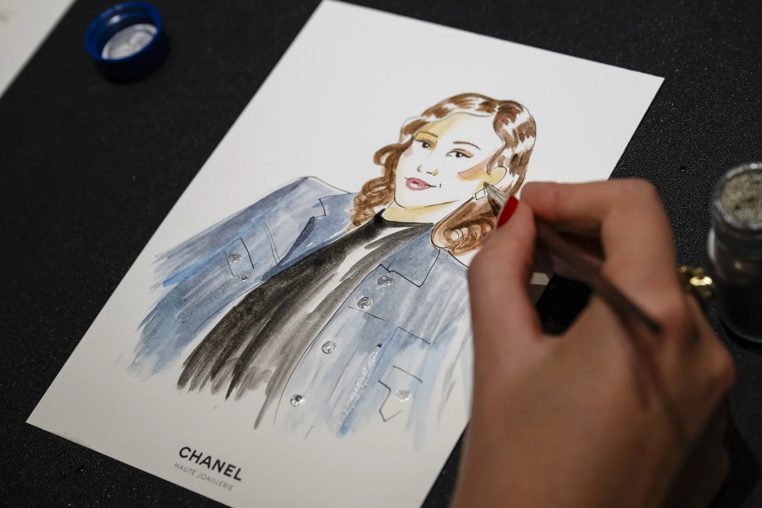 Une personne peint une illustration de style aquarelle d'une femme avec des cheveux bruns, portant un blazer gris et un haut noir. La peinture est une représentation artistique de la femme, avec des détails sur le visage et la coiffure. La pose de la main indique que l'artiste est en train de peindre.