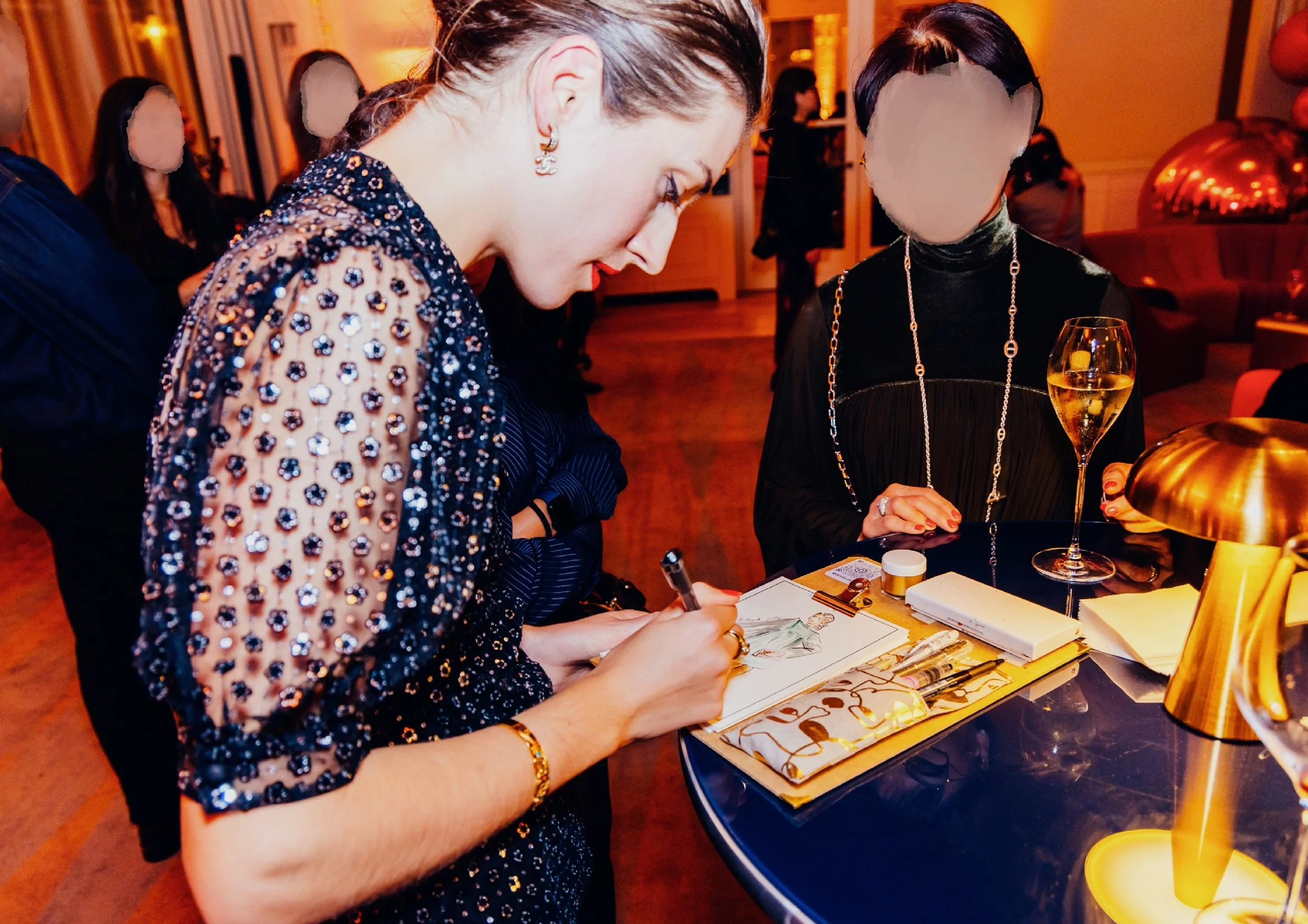 Deux femmes habillées élégamment en train de signer un livre ou un document sur une table lors d'une réception ou d'un événement. L'une d'elles porte une robe ornée de paillettes, l'autre un vêtement noir avec des accessoires en or et un verre de champagne à proximité.