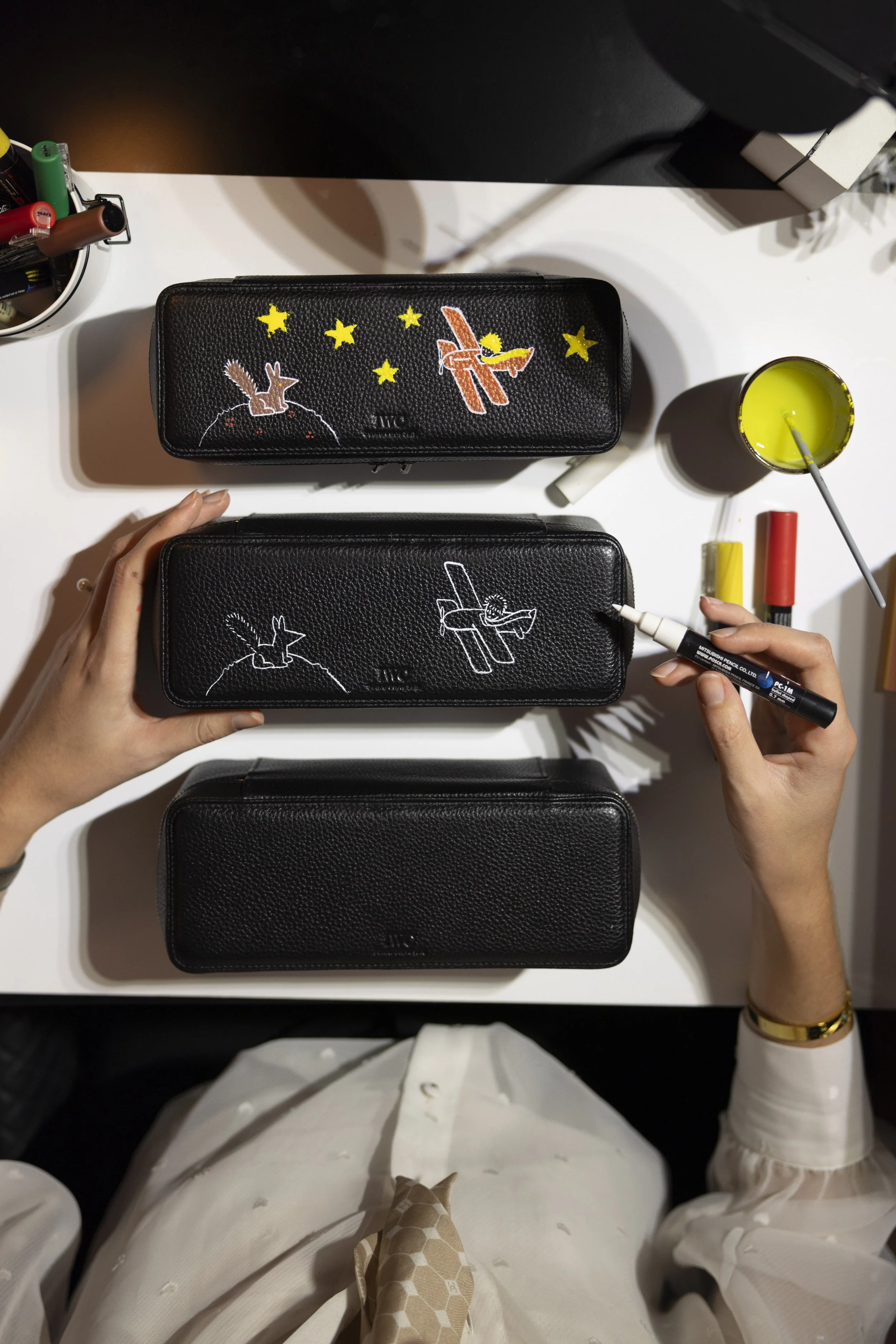 Une personne utilise un stylo pour ajouter un dessin à une des trois étuis noirs en cuir, tous ornés d'un dessin d'oiseau, sur une table blanche. À côté, il y a un contenant avec des stylos, un verre avec une boisson verte, et des blocs-notes.