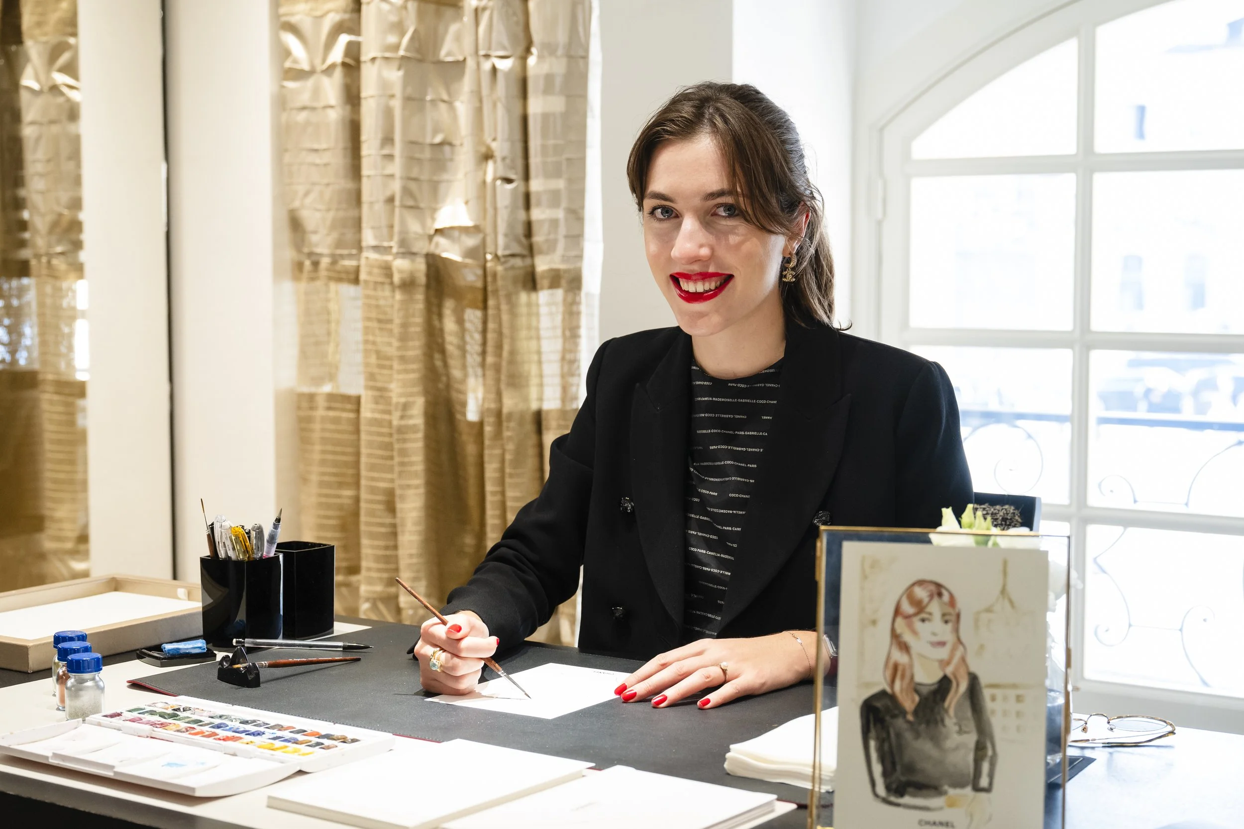 Une femme souriante avec des cheveux bruns, portant un blazer noir et du rouge à lèvres rouge, assise à un bureau avec des pinceaux, des aquarelles, et un tableau représentant une femme avec des cheveux roux. La pièce est bien éclairée avec une grande fenêtre en arrière-plan.