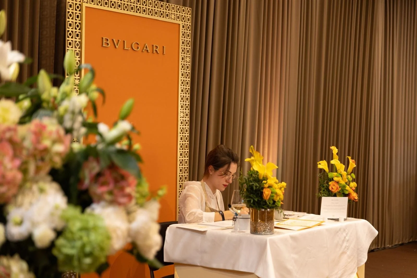 Une femme assise à une table en damier blanc, à côté d'arrangements floraux jaunes et oranges, dans une salle élégante avec des rideaux en tissu marron et un panneau Bvlgari en arrière-plan.