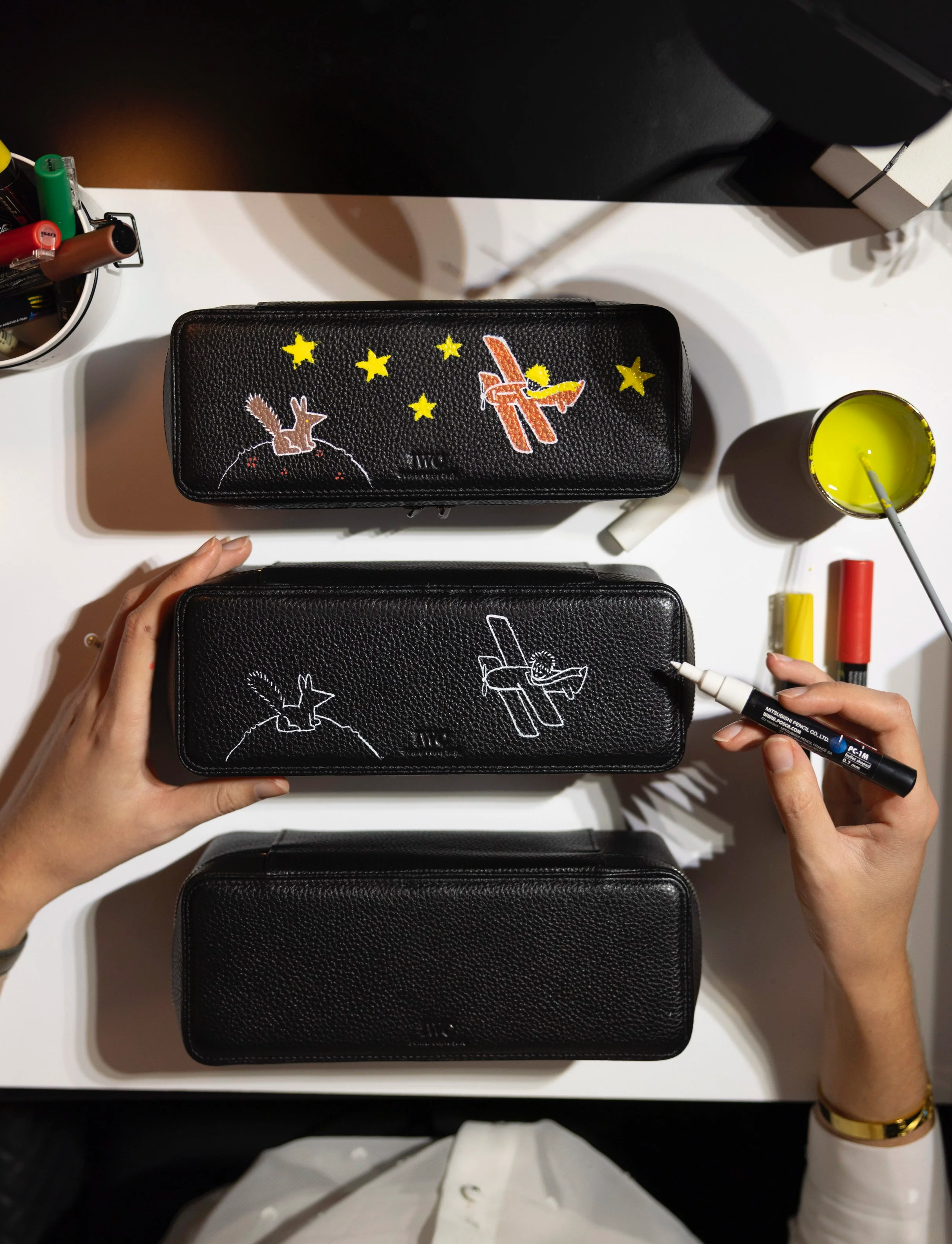 Deux étuis à crayons en cuir noir, posé sur une table blanche, un étant décoré avec un dessin d'avion et de lapin sous des étoiles, l'autre étant partiellement décoré, une main dessinant un lapin avec un marqueur blanc.