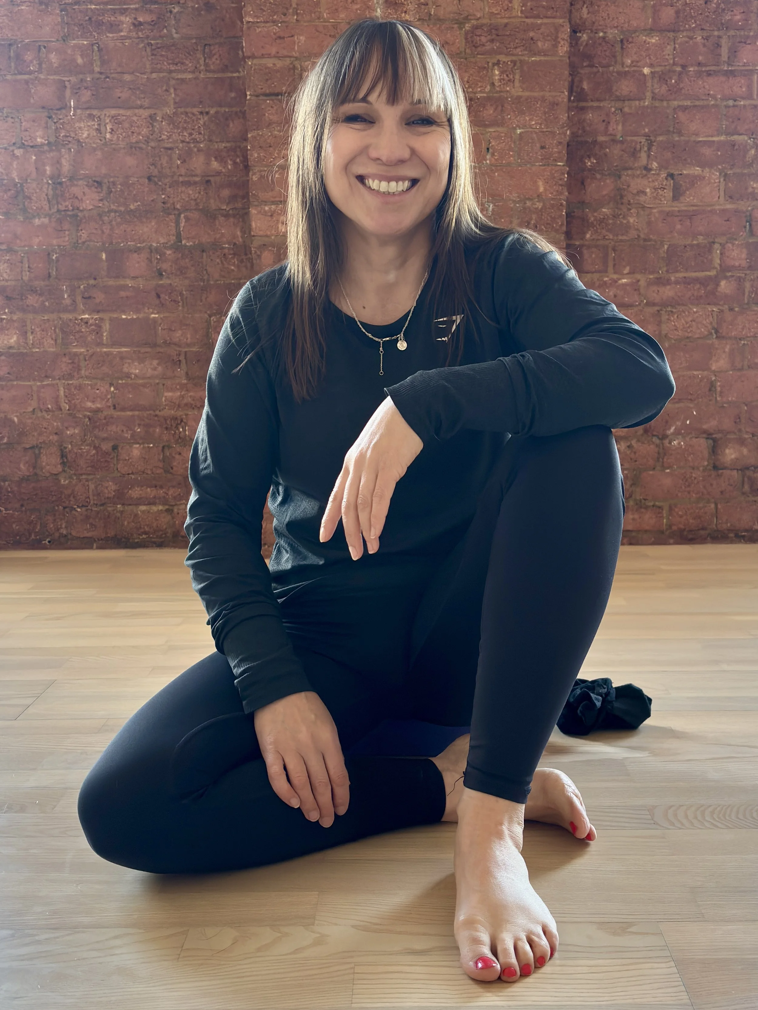 A woman in black athletic wear practicing yoga on a wooden floor in front of a brick wall, sitting in a cross-legged pose with one hand on her knee and the other on her ankle, smiling.