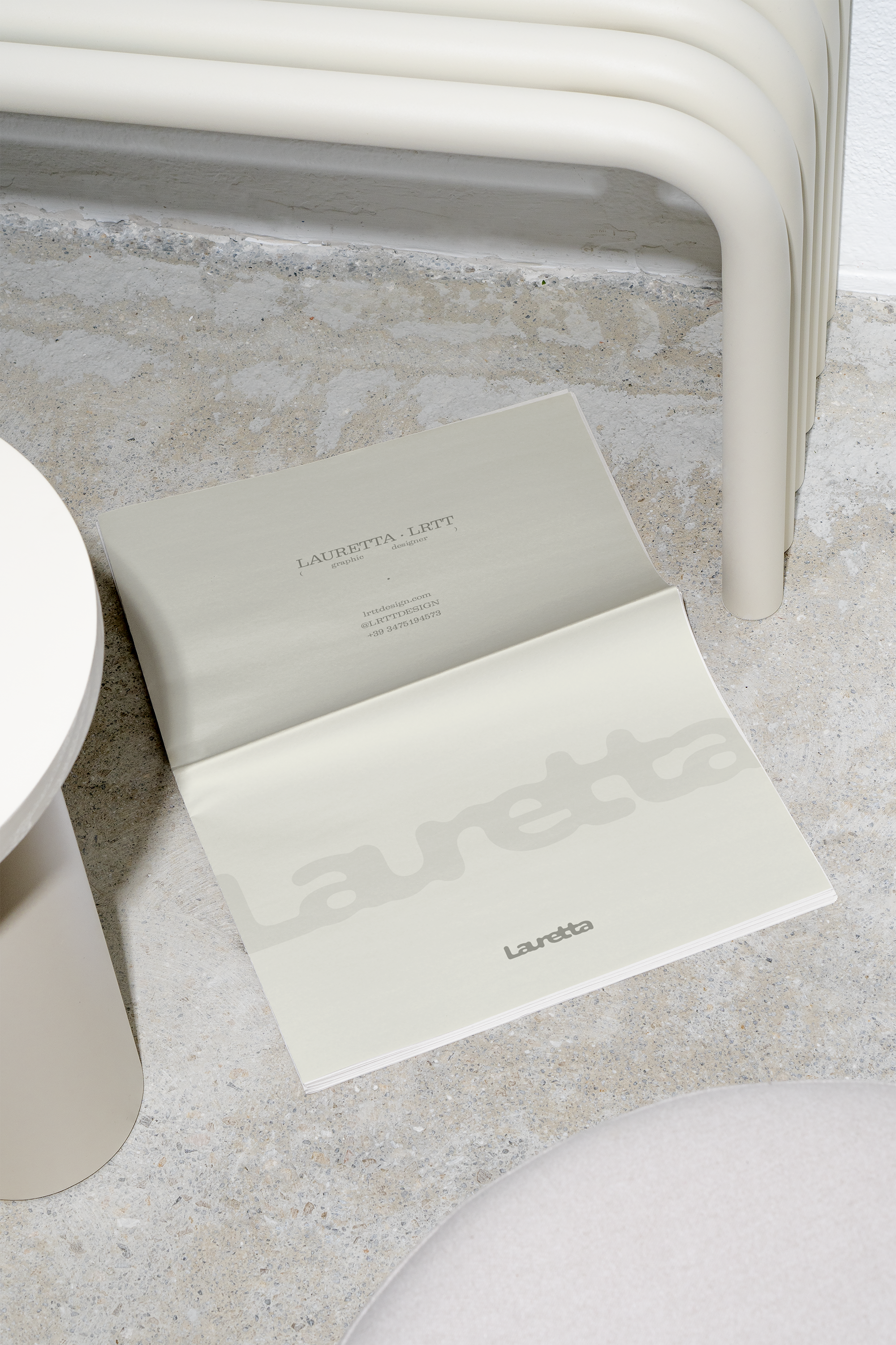 Newspaper con il logo Logo Lauretta | LRTT Design e informazioni di contatto, posizionato su una superficie in cemento, vicino a sedie di colore beige