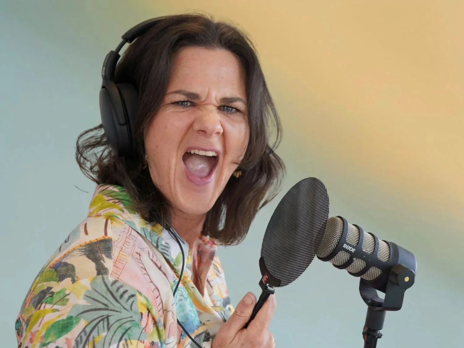 Podcast host and executive coach Penny Terry wearing headphones and a colourful shirt, holding a podcast microphone and speaking.
