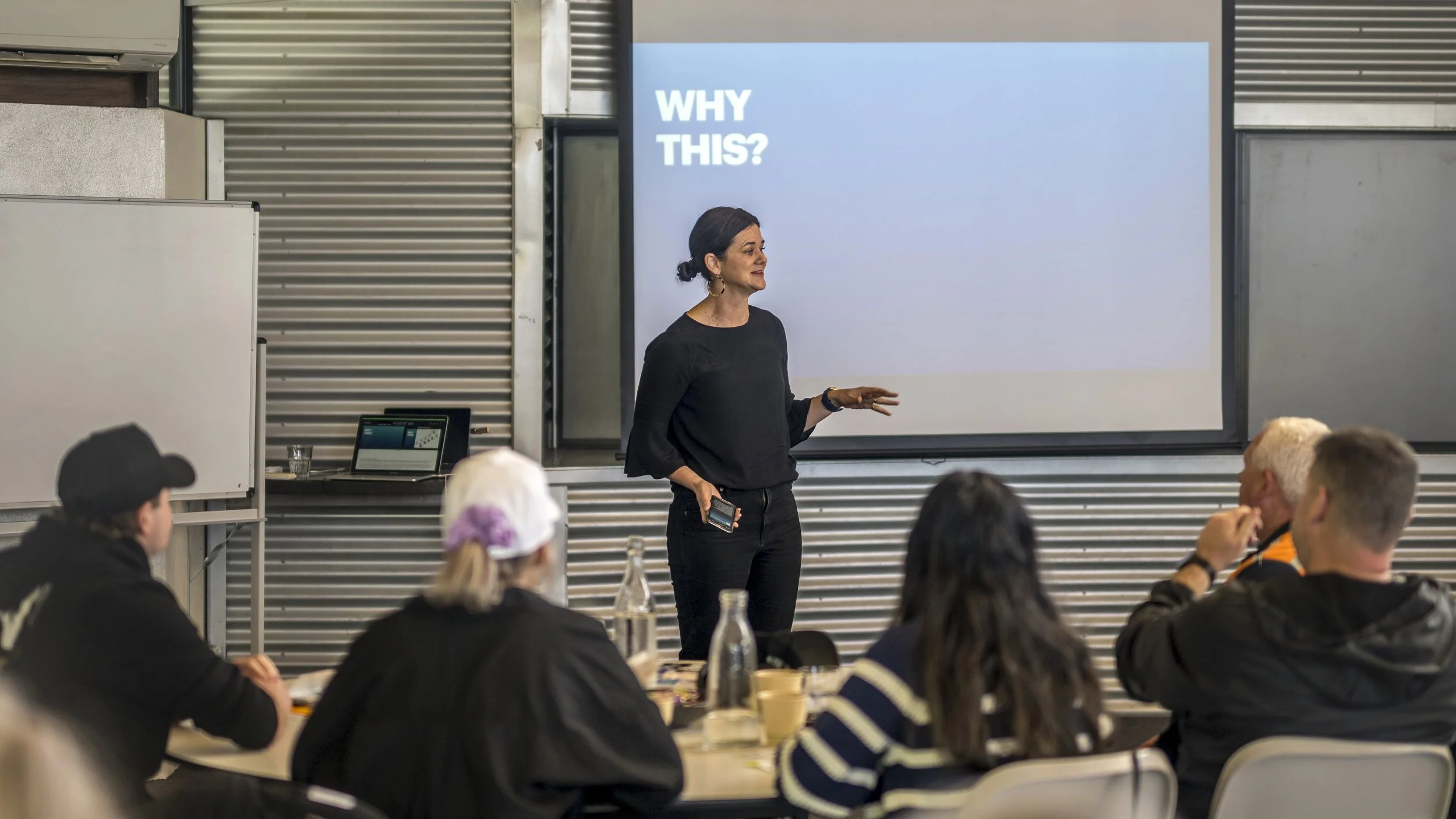 Penny Terry at the front of a training room, presenting to a team, with a large screen displaying the words "WHY THIS?"