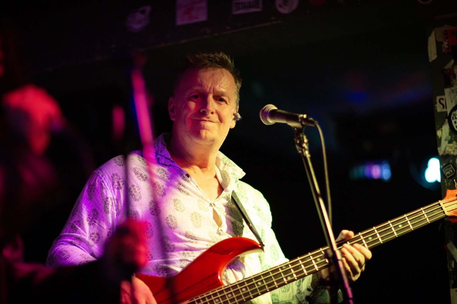 A man playing a bass guitar on stage with a microphone in front of him, illuminated by colorful stage lights, wearing a patterned button-up shirt.