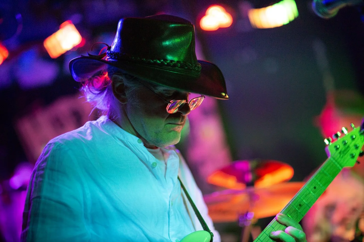 A man wearing a cowboy hat and glasses playing an electric guitar in a dimly lit, colorful environment with purple, blue, and green lights.