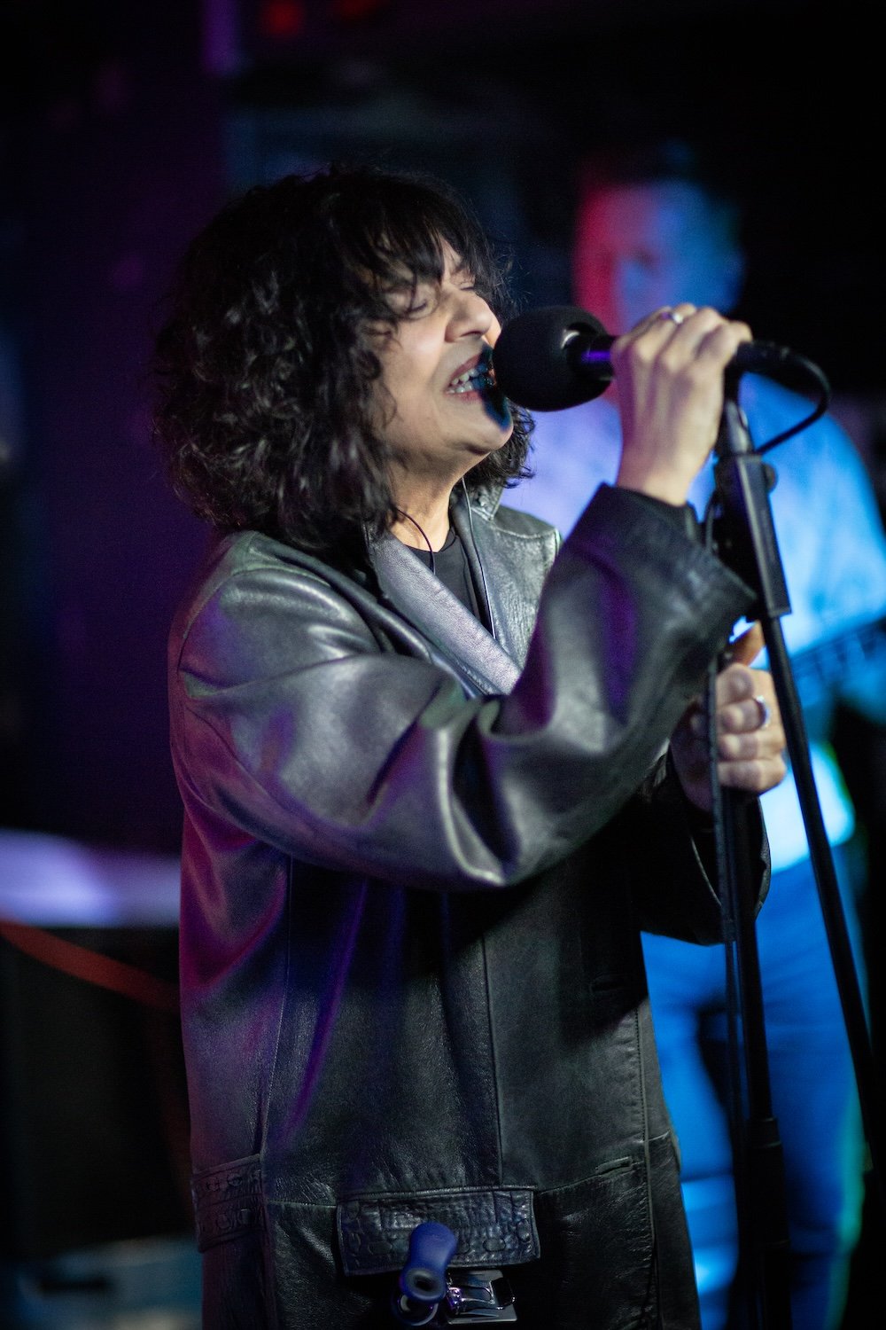 A woman with curly black hair singing into a microphone on stage, wearing a black leather jacket, in a dimly lit setting with colorful lights.