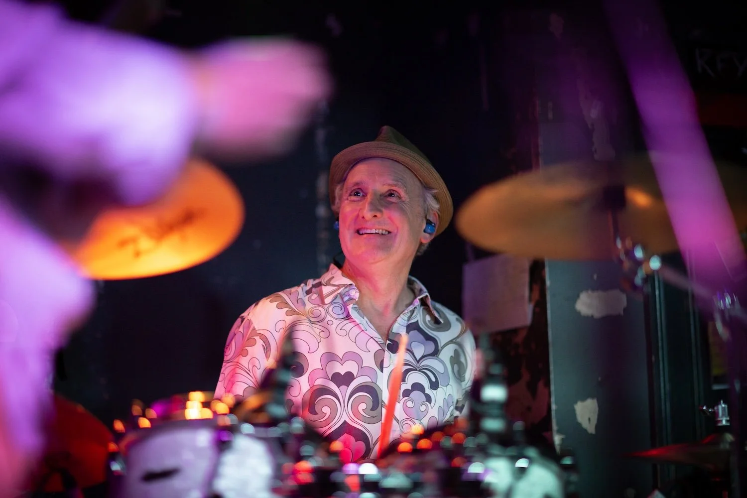 A smiling man with gray hair wearing a hat and patterned shirt, playing drums on stage under colorful lights.