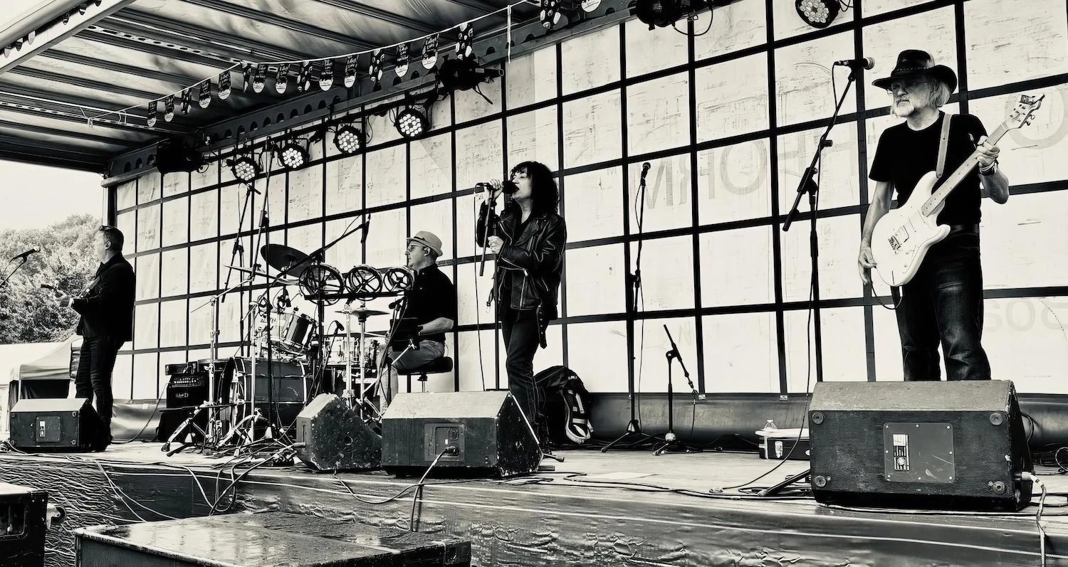 A four-member band performing on an outdoor stage with instruments including a guitar, drums, and microphone, in black and white.