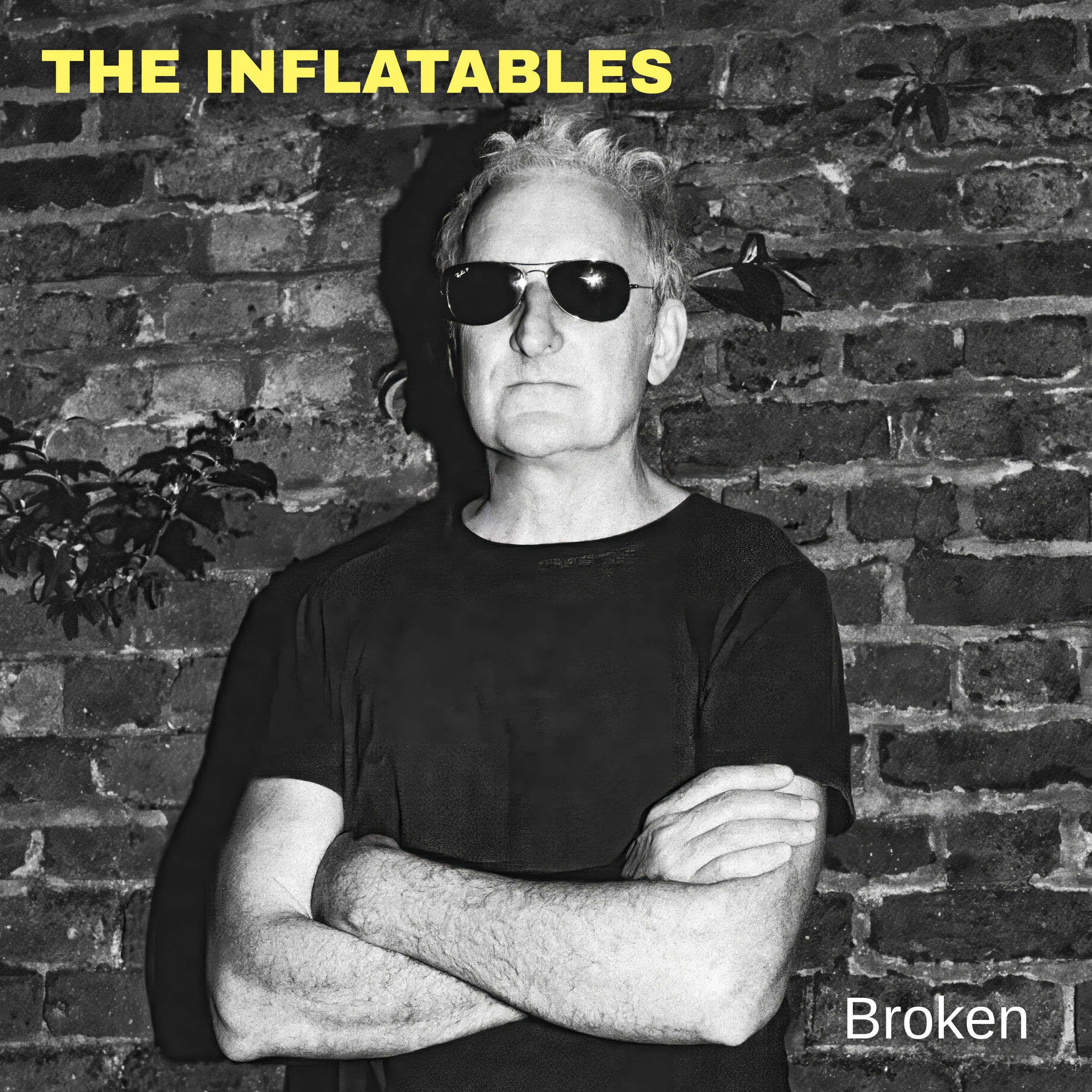 Black and white photo of The Inflatables drummer, Sam Phillips, with sunglasses, arms crossed, standing in front of a brick wall. The words "THE INFLATABLES" are at the top in yellow text, and the word "Broken" is at the bottom right in white text.