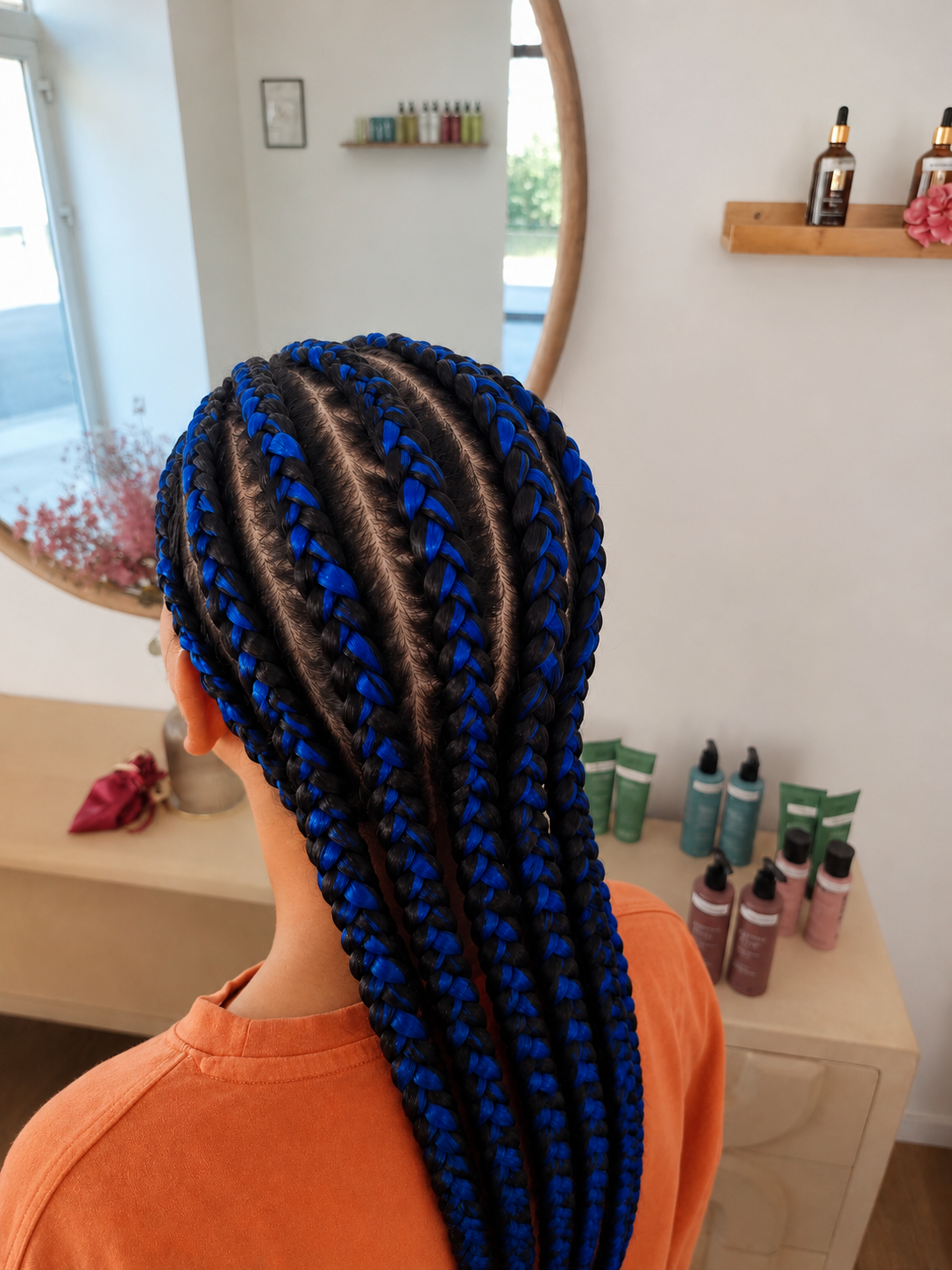 Coiffure à tresses avec des fils bleus et noirs, vue de l'arrière d'une personne dans un salon de coiffure. En arrière-plan, un miroir, des produits capillaires sur une étagère, et un meuble avec des bouteilles de shampoing ou de soins capillaires.