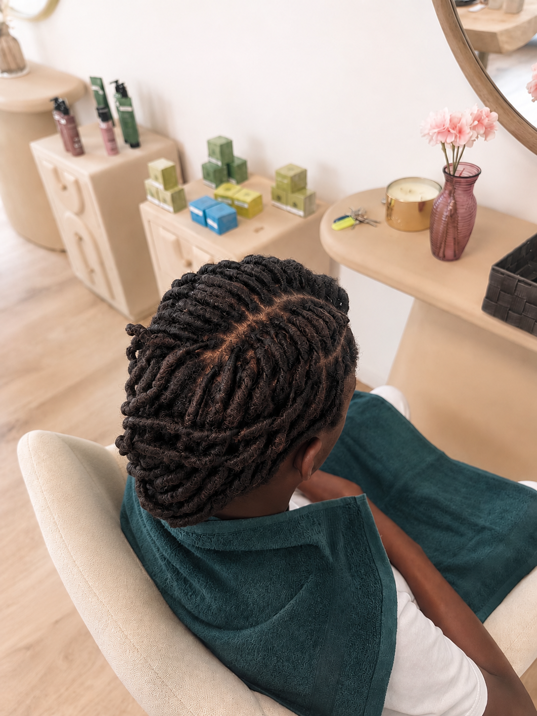 Personne assise dans un fauteuil, coiffée de dreadlocks, dans un salon ou un spa, avec des produits capillaires sur une console derrière.
