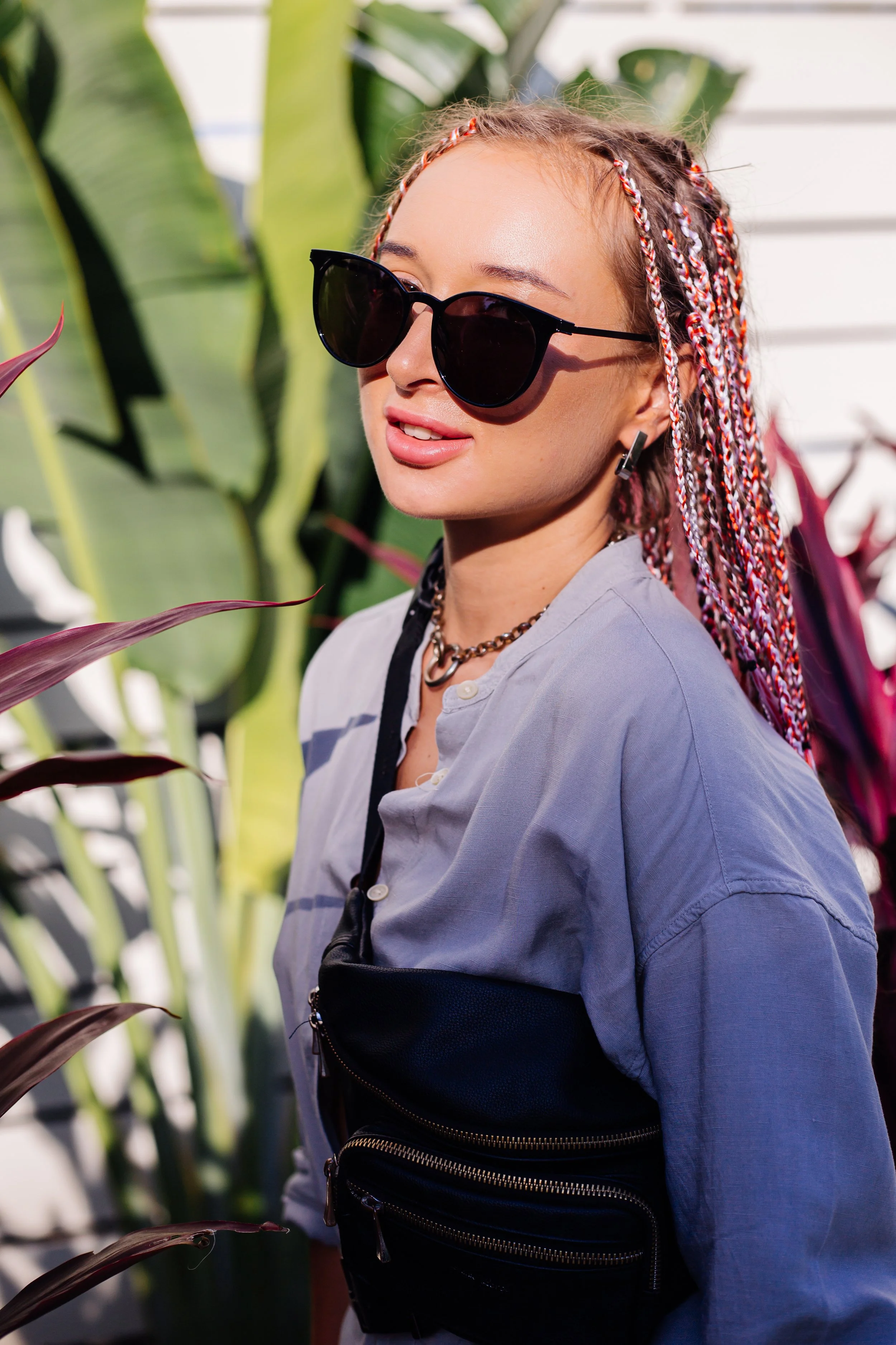 Jeune femme avec des lunettes noires et une chemise grise, portant un sac banane noir, posant à l'extérieur parmi des plantes tropicales.