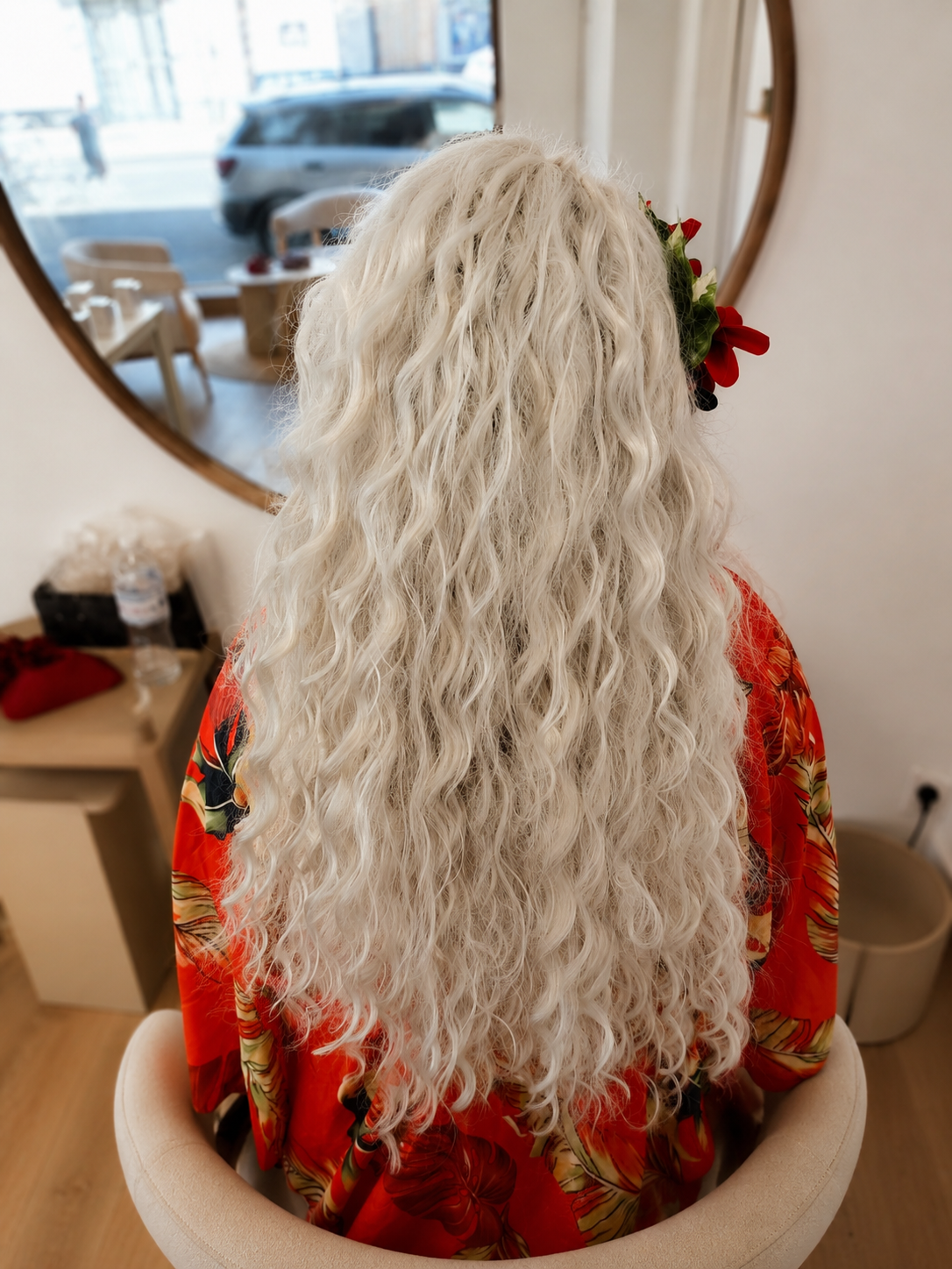 Une personne avec de longs cheveux blonds, ondulés, assise dans un salon. Elle porte une robe rouge avec des motifs floraux et a des fleurs rouges dans les cheveux.