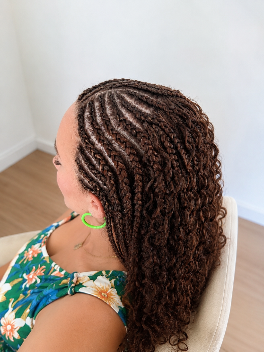 Une femme aux cheveux bouclés bruns avec de nombreuses tresses laquées, portant un haut floral, une boucle d'oreille verte et une chaîne fine, assise sur une chaise blanche dans une pièce claire.