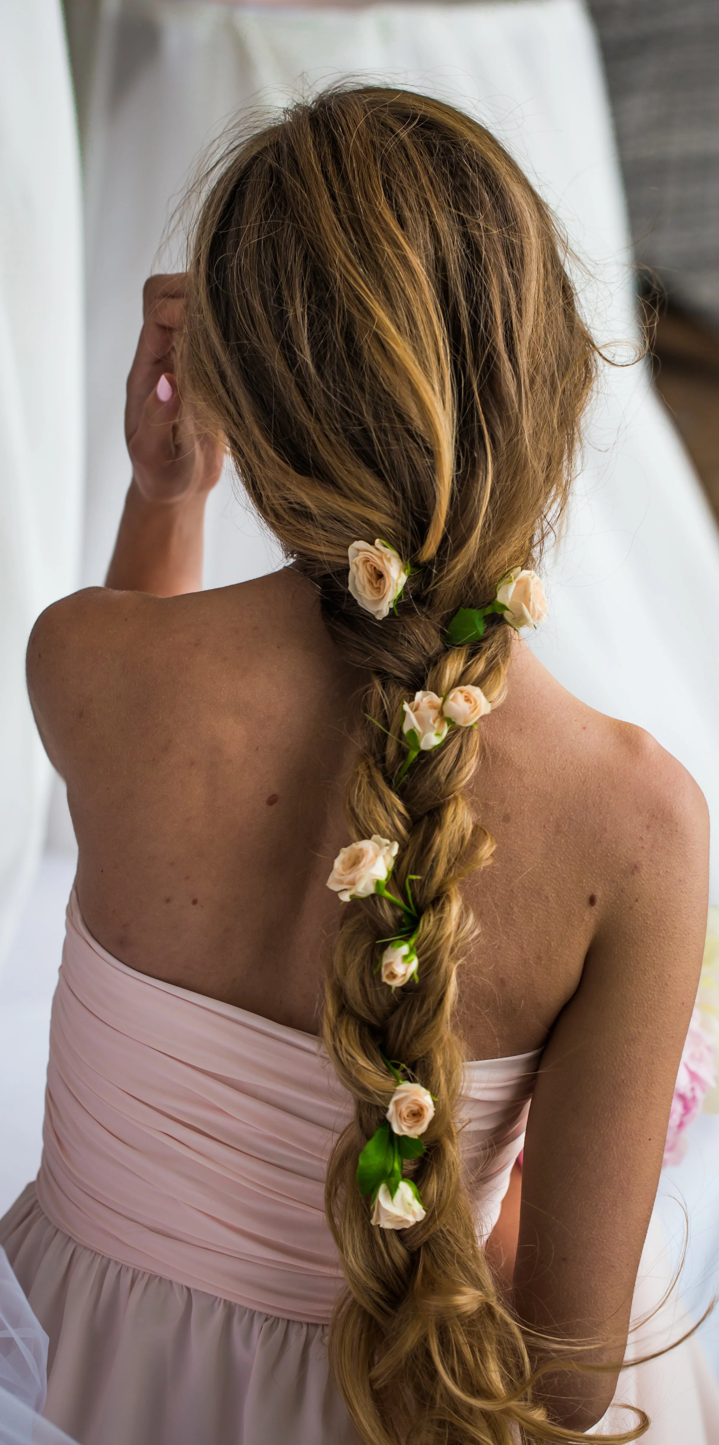 Cheveux longs, blonds, en tresse ornée de petites roses blanches et vertes, vue de derrière, portant une robe rose pâle à épaule nue.