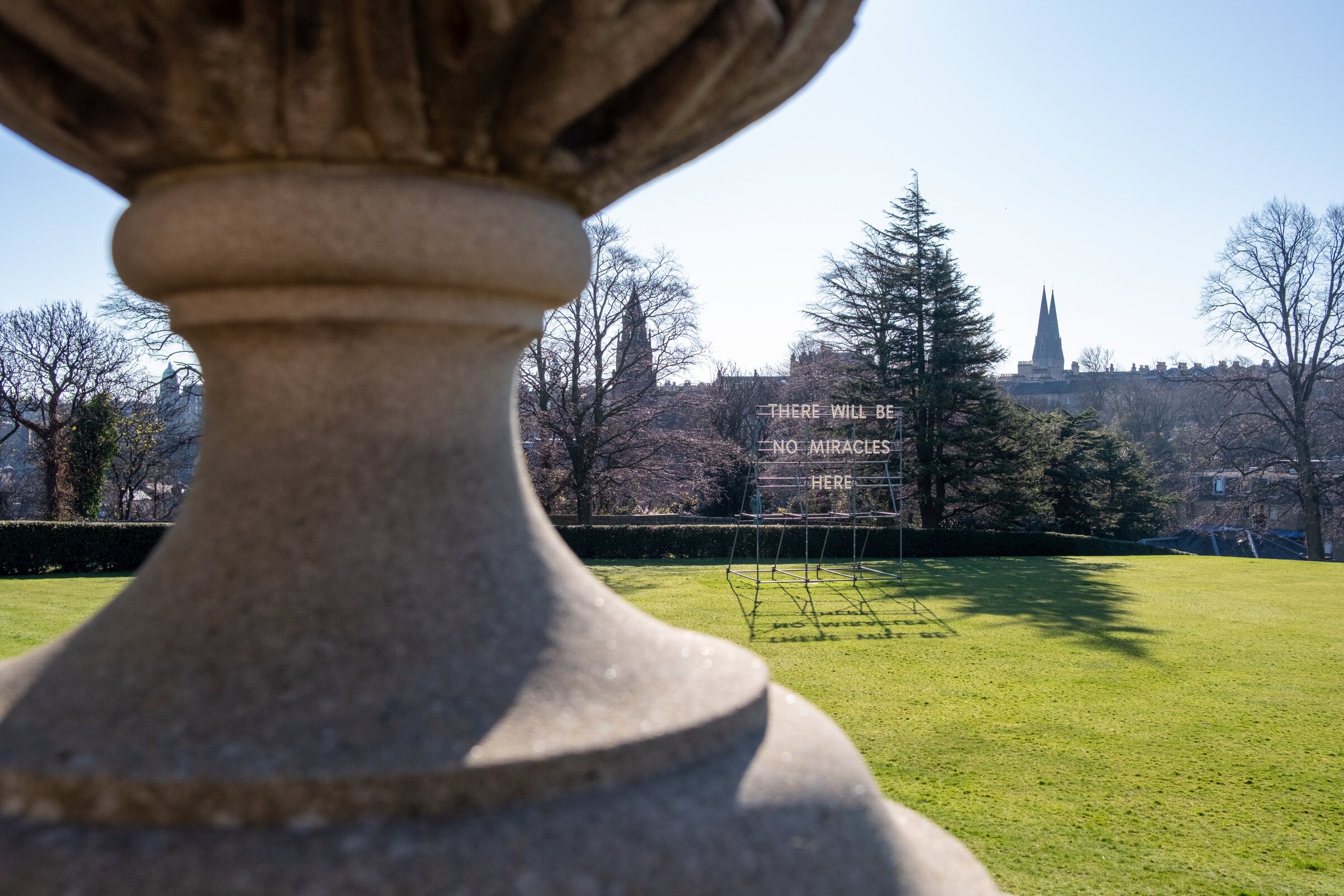 Illuminated white letters reading 'There Will Be No Miracles Here' beside a stone garden urn