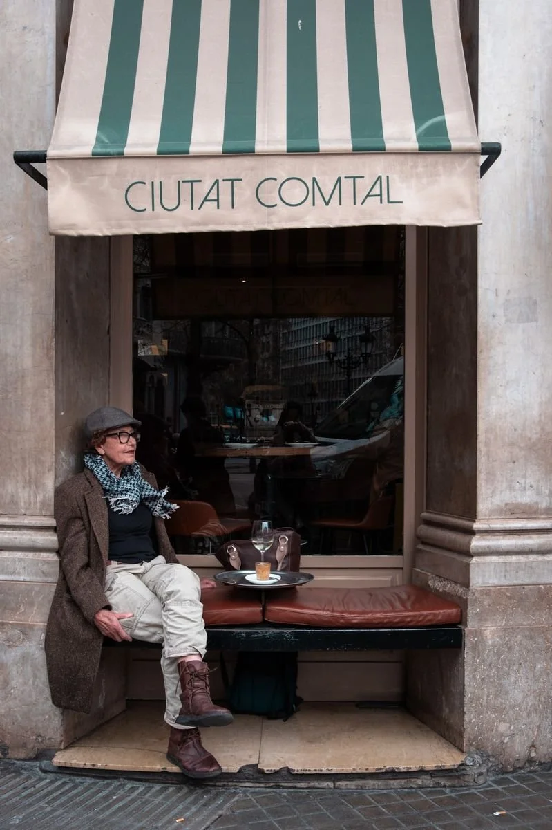 An elderly woman relaxes at an outdoor cafe table beneath the Ciutat Comtal awning