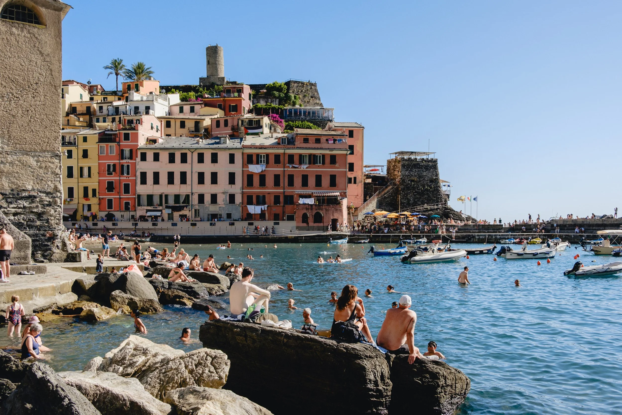 People swimming and relaxing by the rocky shoreline of a harbor with colorful buildings and boats, in a sunny seaside town.