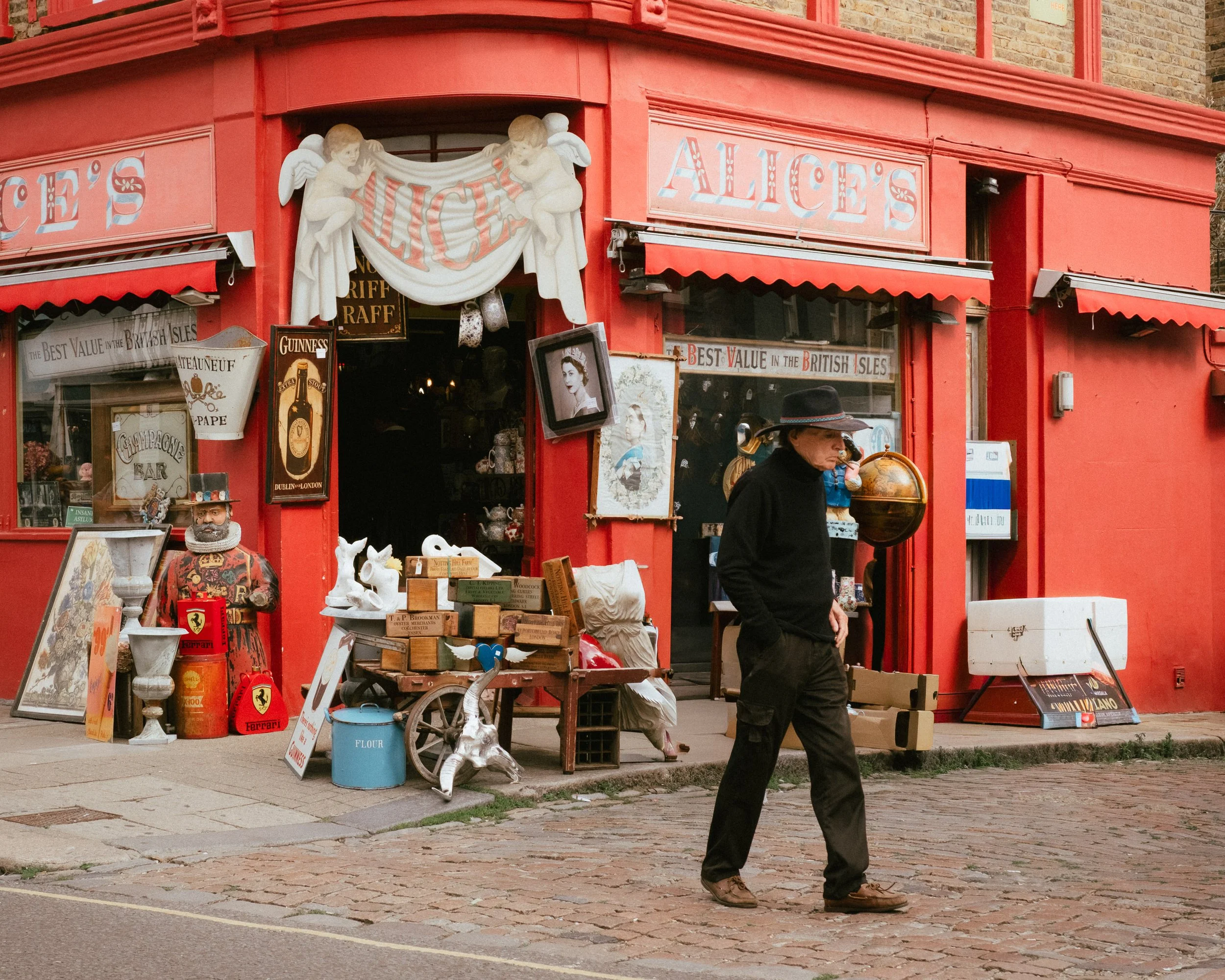 A person in black clothing and a wide-brimmed hat walking past a red antique shop called Alice's. The shop has vintage items outside, including books, a horse statue, and various decorative objects.