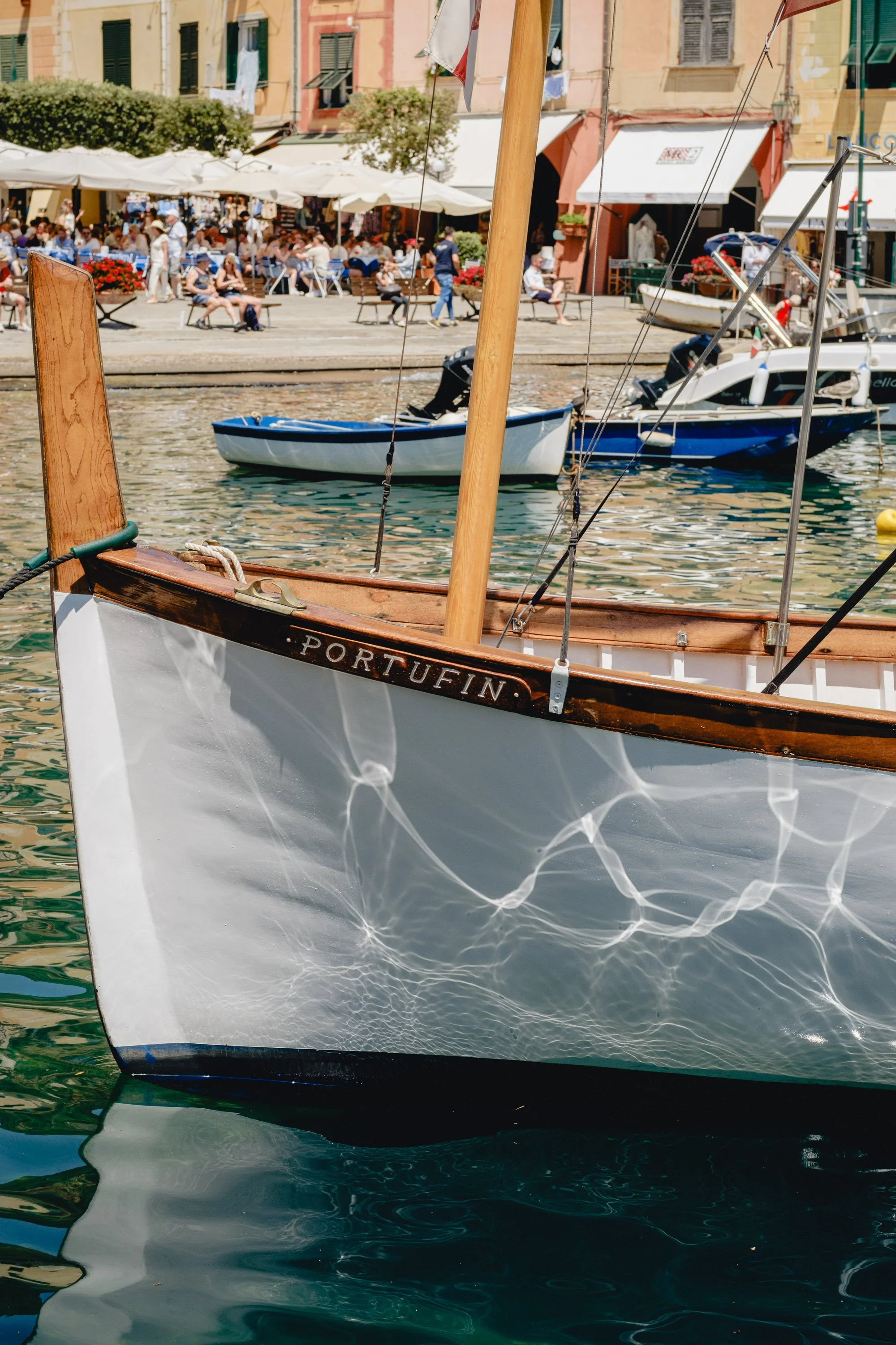 Close-up of a wooden sailboat named 'Portufin' docked in a harbor, with a lively outdoor cafe and people seated at tables along a waterfront in the background.
