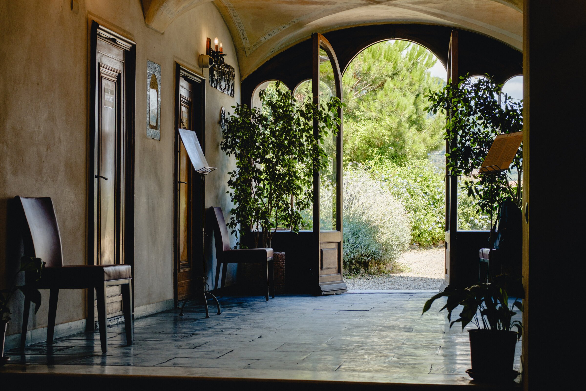 A shaded stone loggia with potted plants opens onto a green garden through arched doorways
