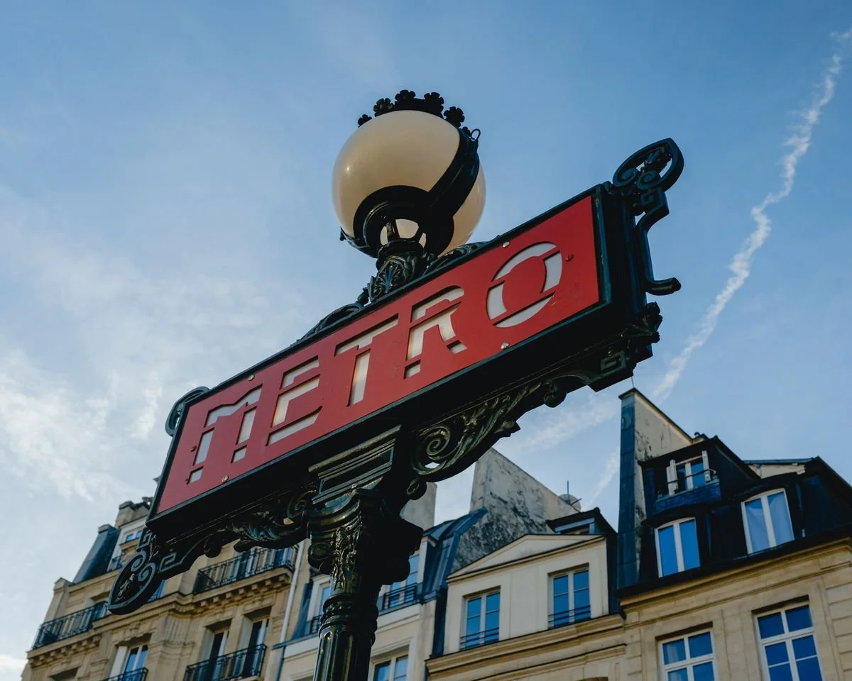 A red enamel Paris Métro sign photographed from below against a pale blue sky