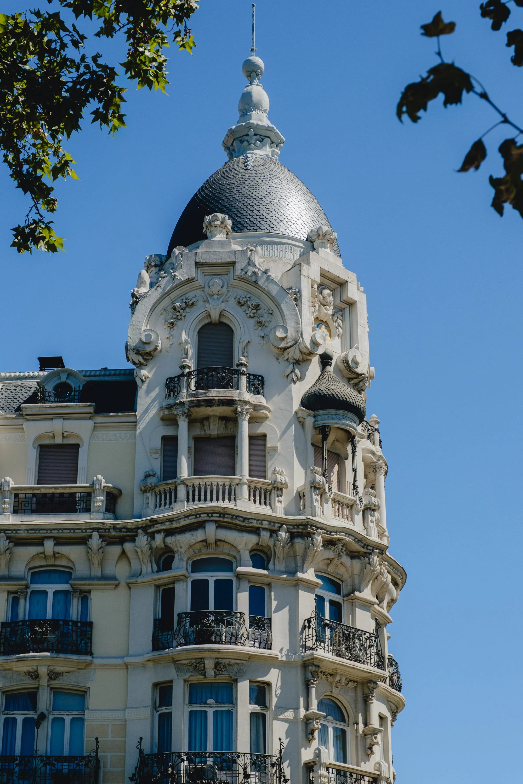 Close-up of ornate historic building with a rounded turret, decorative sculptures, balconies with wrought iron railings, and a domed rooftop against a blue sky, framed by tree leaves.