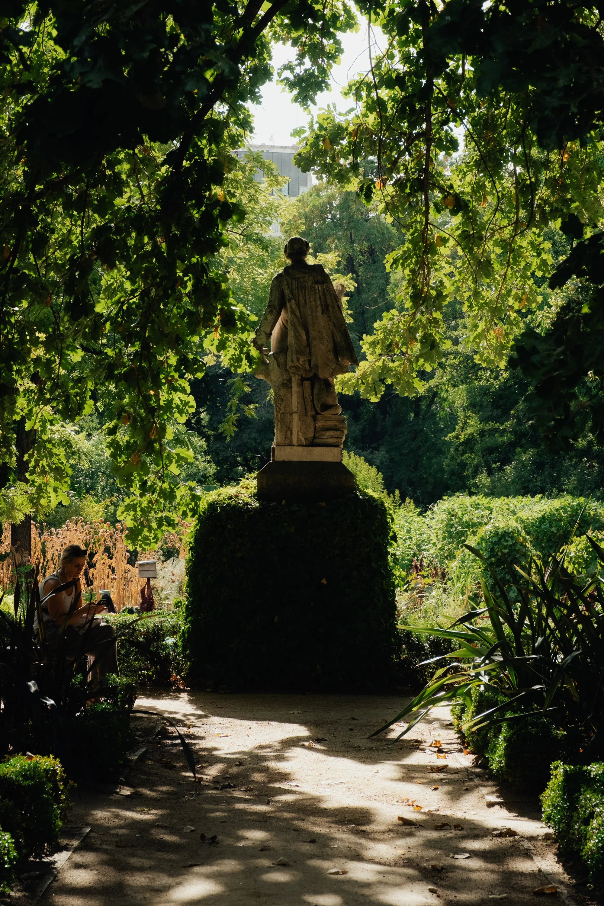 A statue of a person standing on a pedestal in a lush green park, framed by leafy trees, with a woman sitting on a bench nearby.