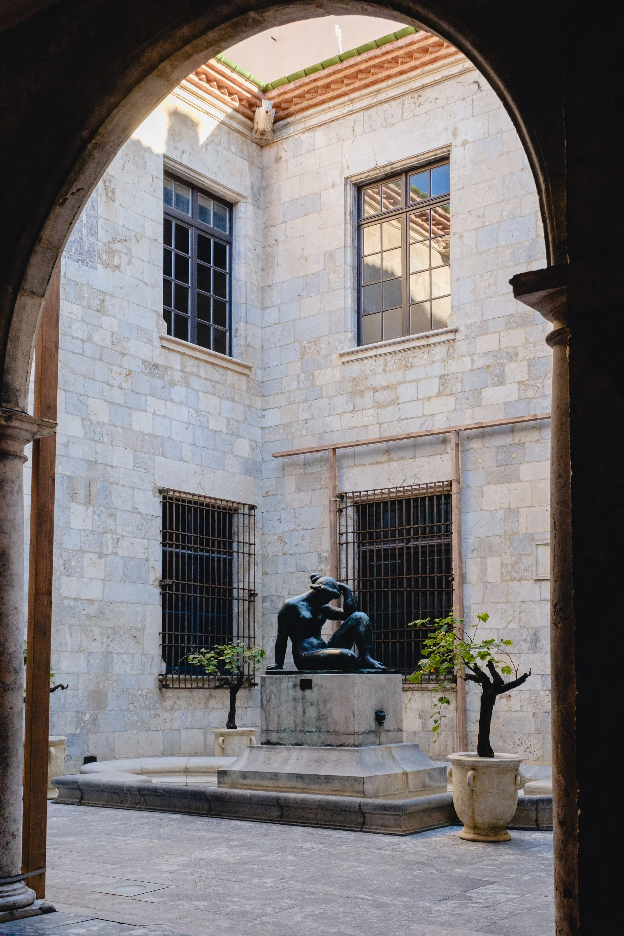 Inner courtyard with a bronze sculpture of a seated woman, two small trees in planters, and barred windows on stone walls, seen through an arched doorway.