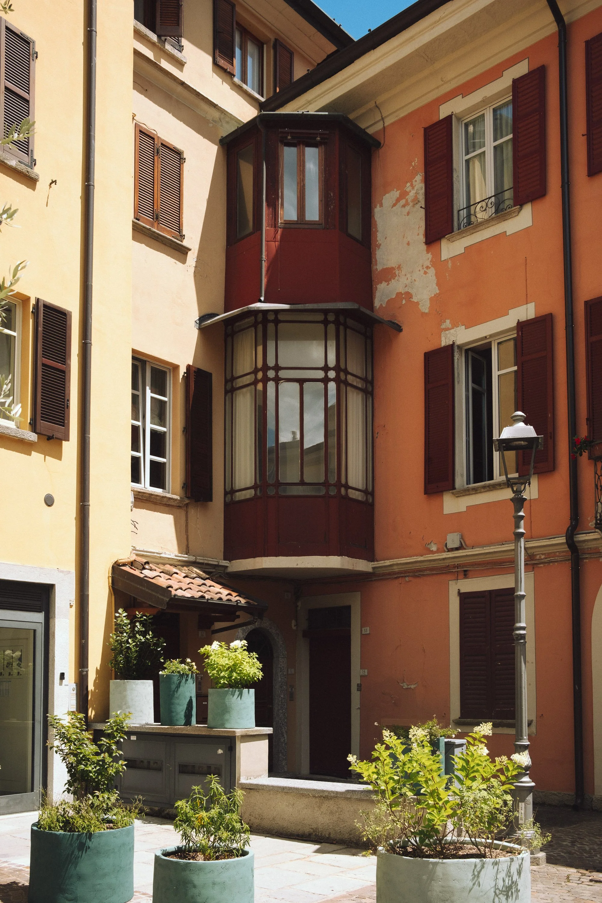 Colorful European-style building with a rounded bay window and open shutters, flowerpots on the sidewalk, and a street lamp.