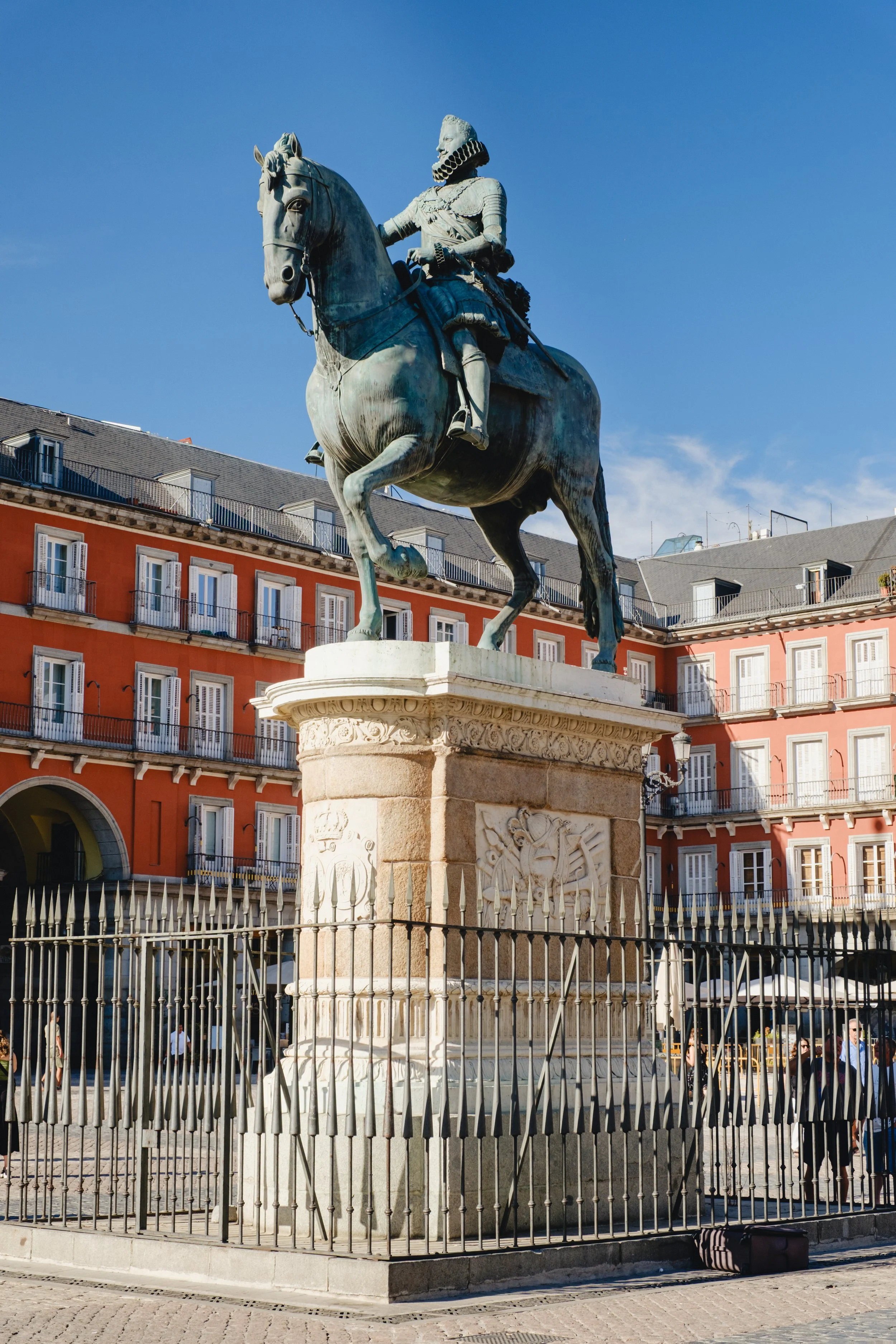 Equestrian statue of a historical figure on horseback in a city square, surrounded by a decorative fence and colorful apartment buildings under a clear blue sky.
