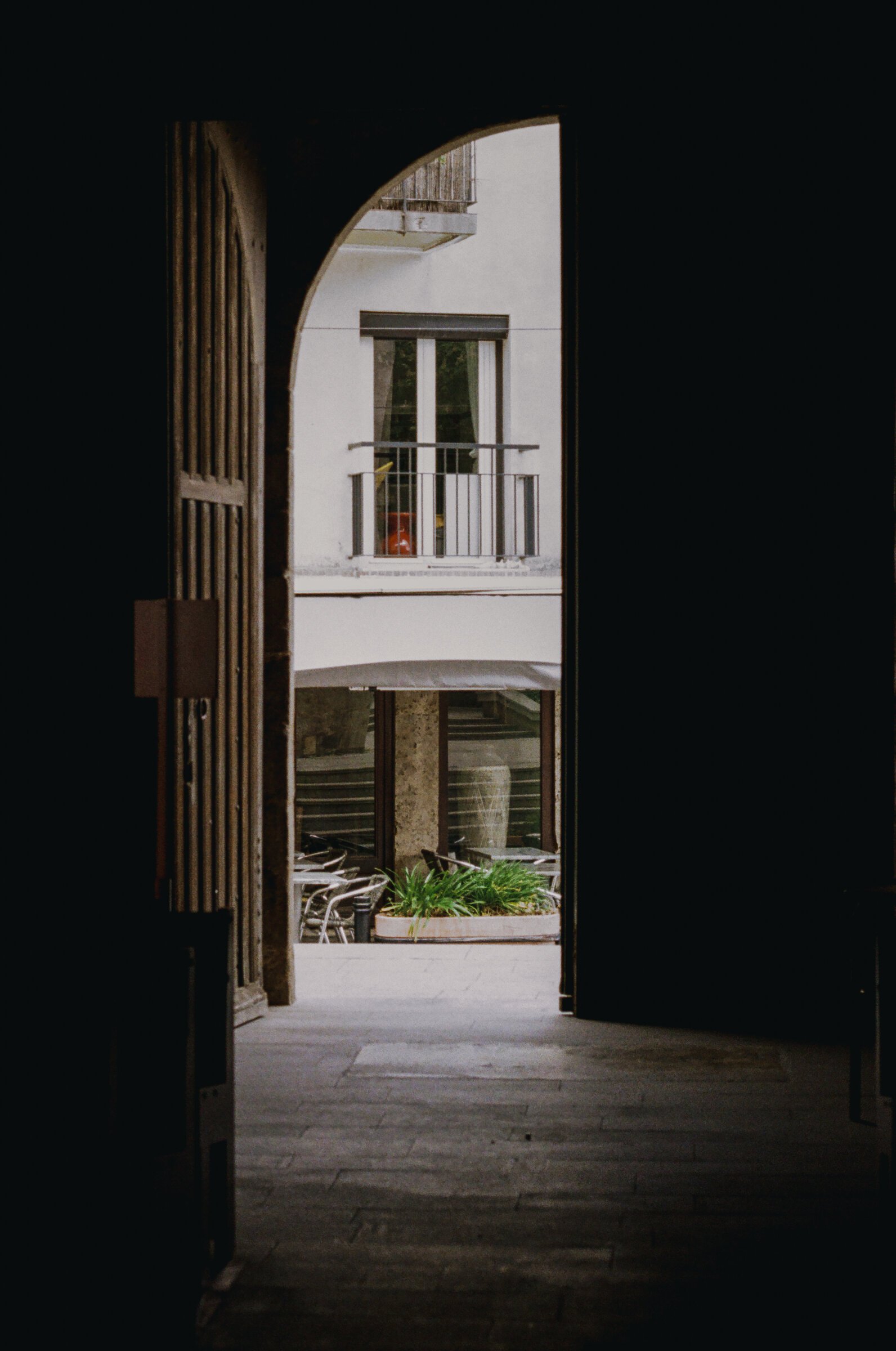 A dark stone passageway opens onto a sunlit Argelès courtyard with café tables and a wrought-iron balcony