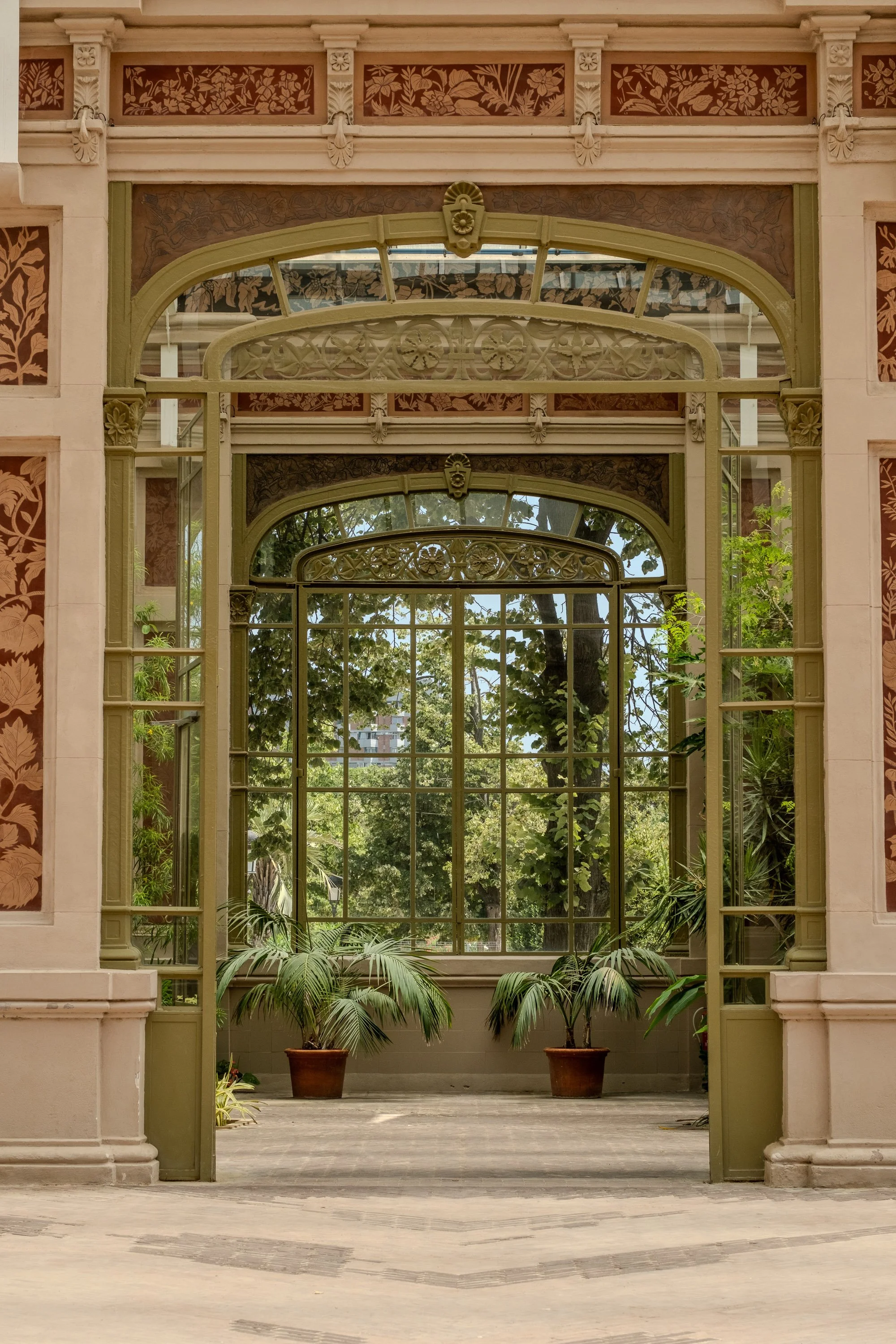 An ornate, vintage-style greenhouse with large windows, decorative metalwork, and potted plants inside. Through the windows, trees and a sunny sky are visible.
