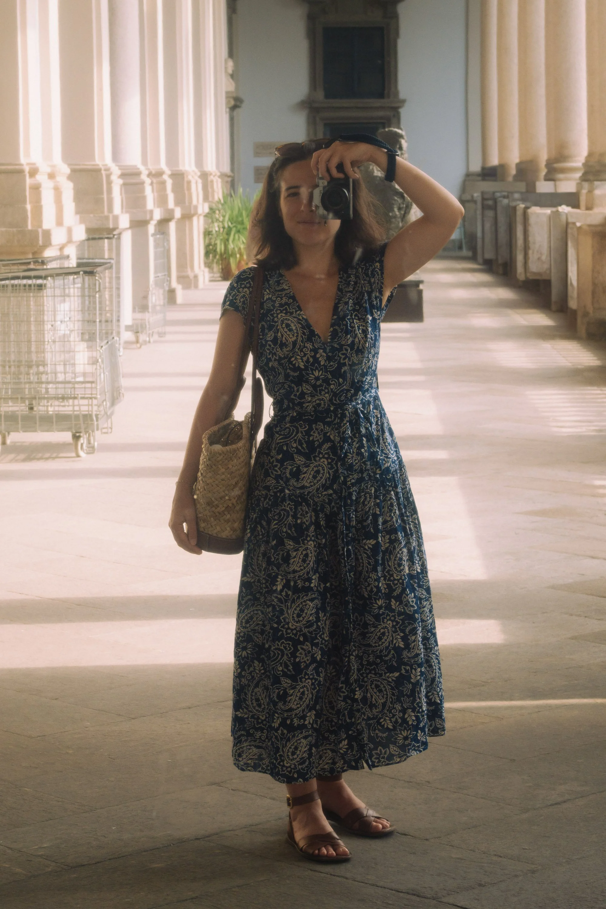 A woman in a blue floral dress taking a selfie in a museum corridor with tall columns and large windows.