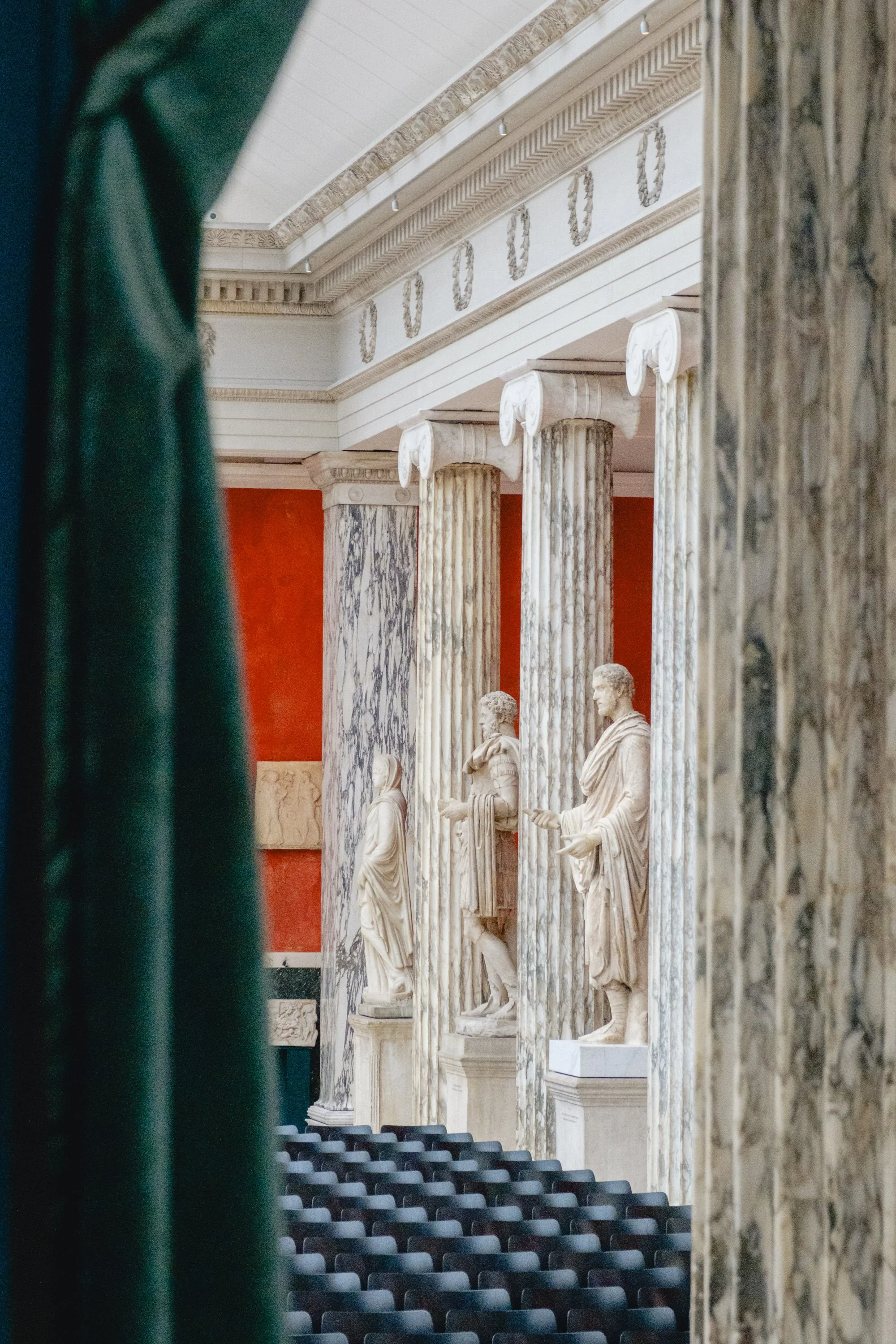 View of Greek or Roman marble statues of historical figures inside a museum or gallery, framed by green curtains, with architectural columns and detailed molding.
