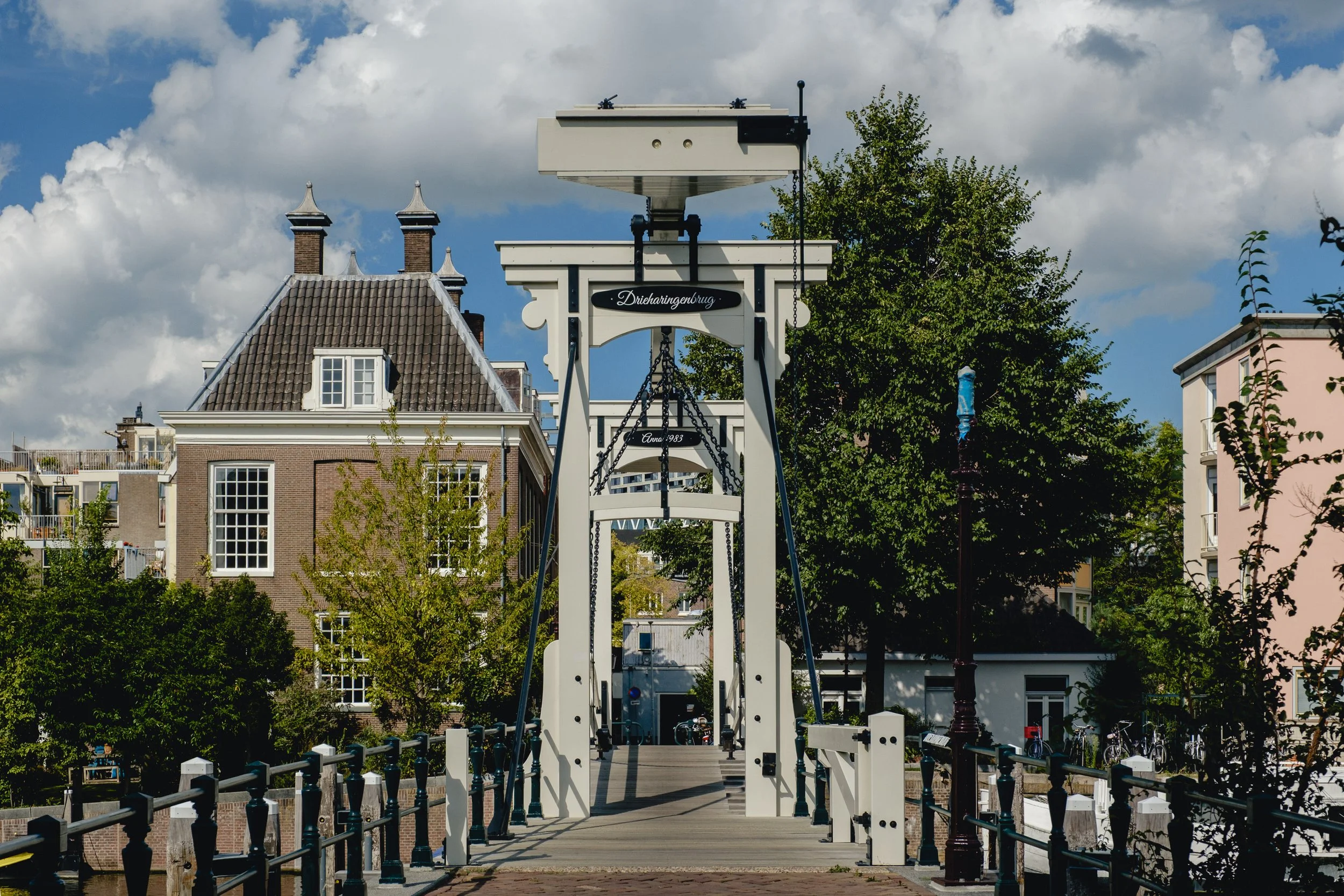 A white drawbridge with black accents in a residential area, with trees and houses in the background, under a partly cloudy sky.