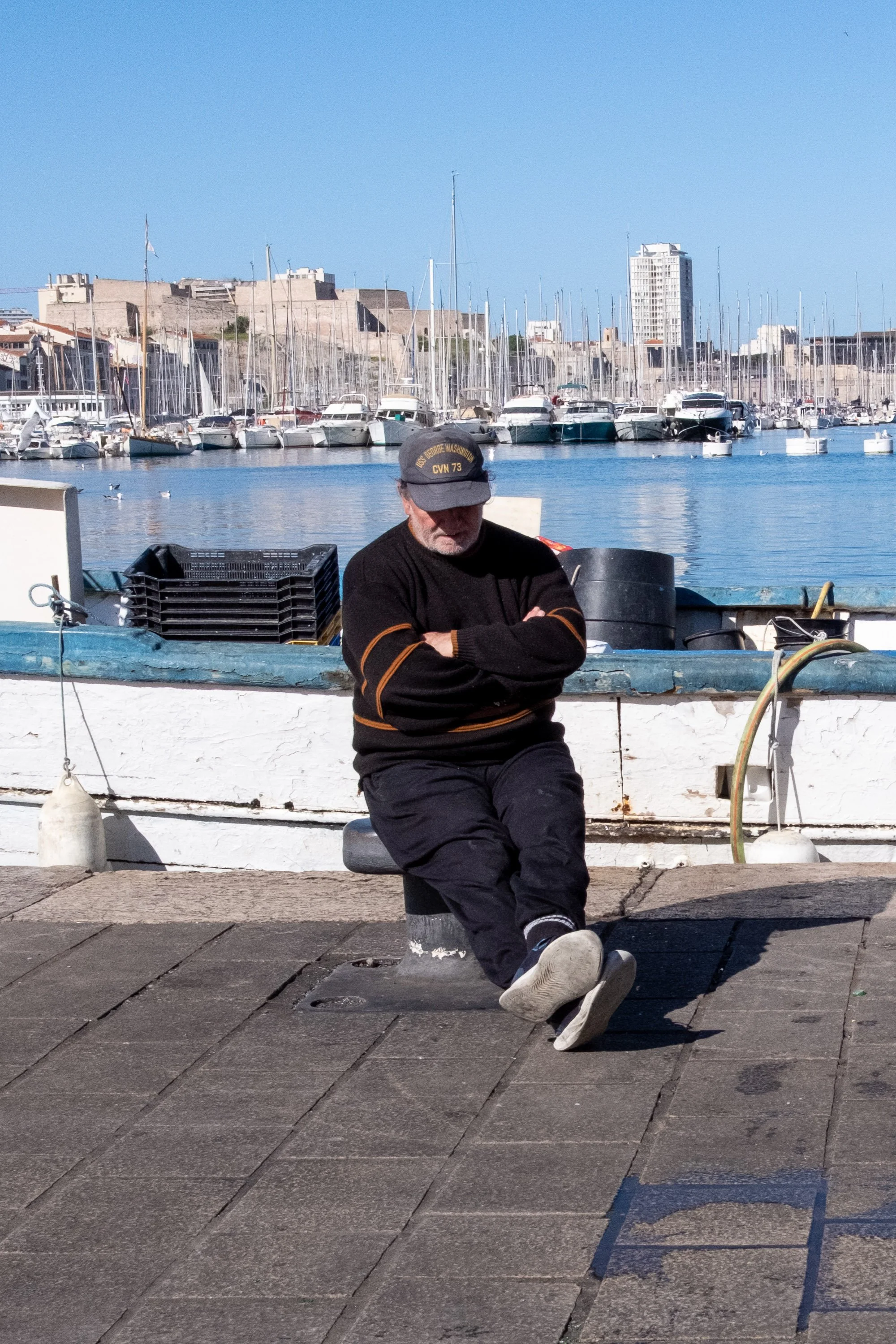 A man sitting on a bench near a marina with yachts, wearing a cap and black clothing, with arms crossed and one leg extended.