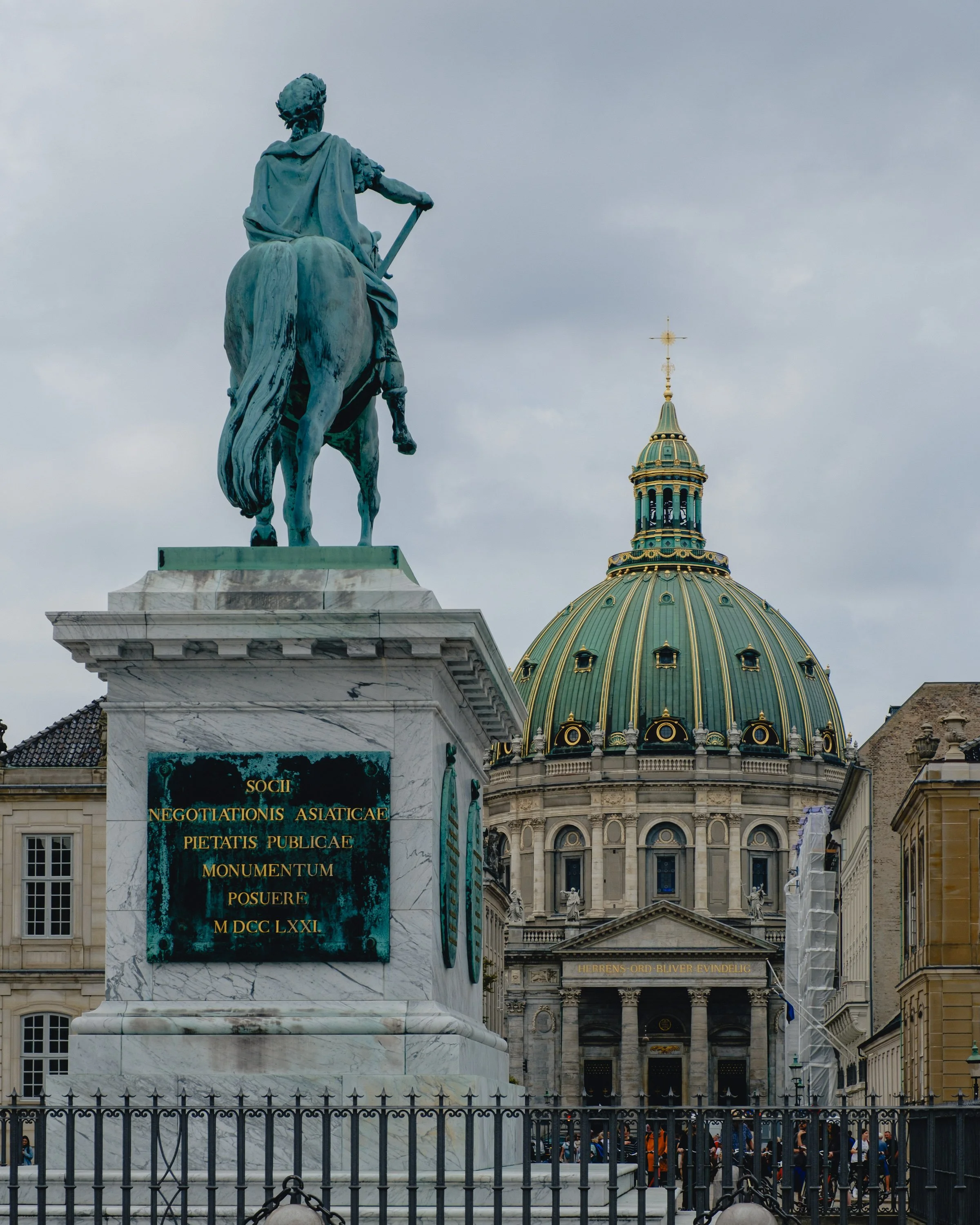 Equestrian statue of a historical figure in front of a large domed building with a gold and green roof in a city square, with a cloudy sky overhead.