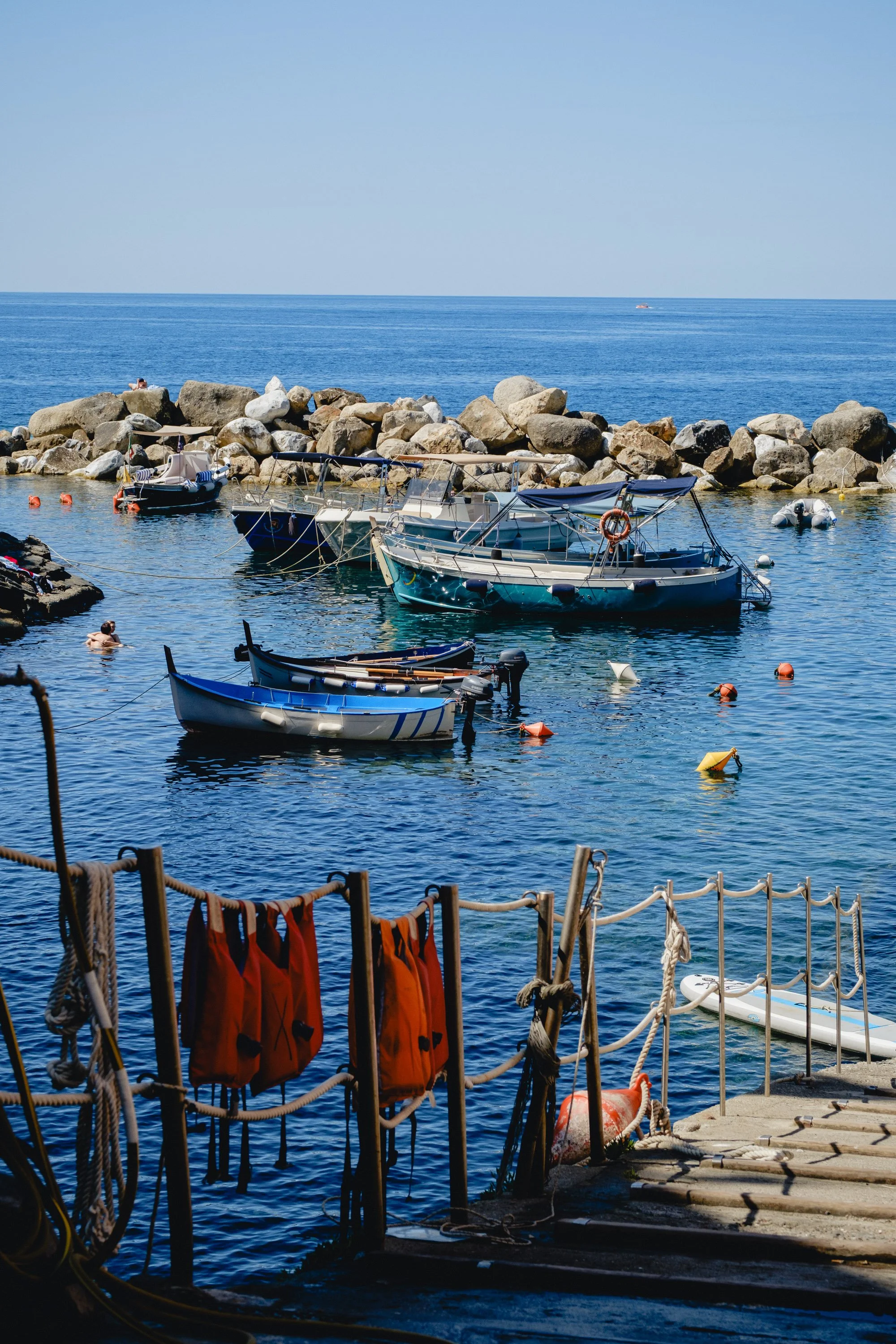 Boats docked in a harbor with rocks and the open sea in the background, life jackets hanging on a rope in the foreground.