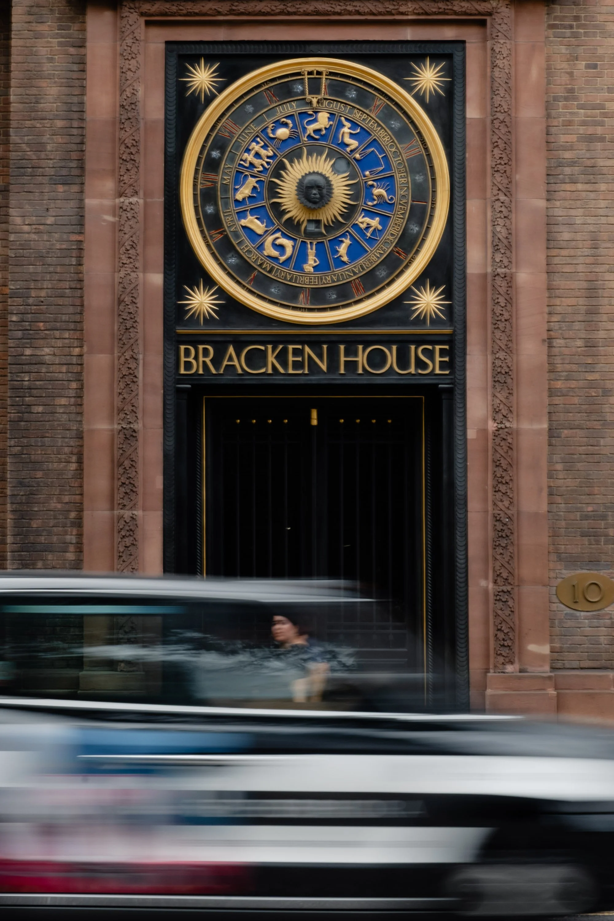 A large ornate clock with zodiac signs and months, mounted above the entrance to Bracken House, with a person walking past and a blurred vehicle in front.