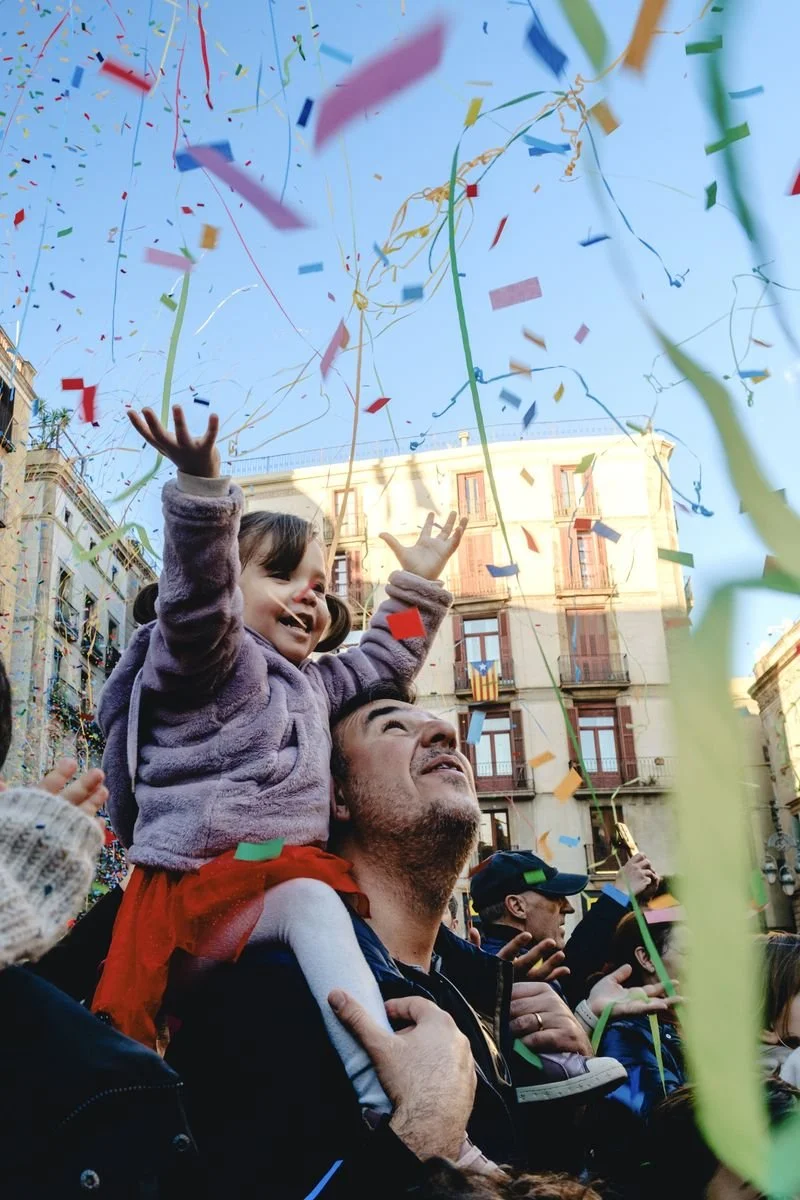 A girl on her father's shoulders reaches up as confetti streamers rain down at a festival